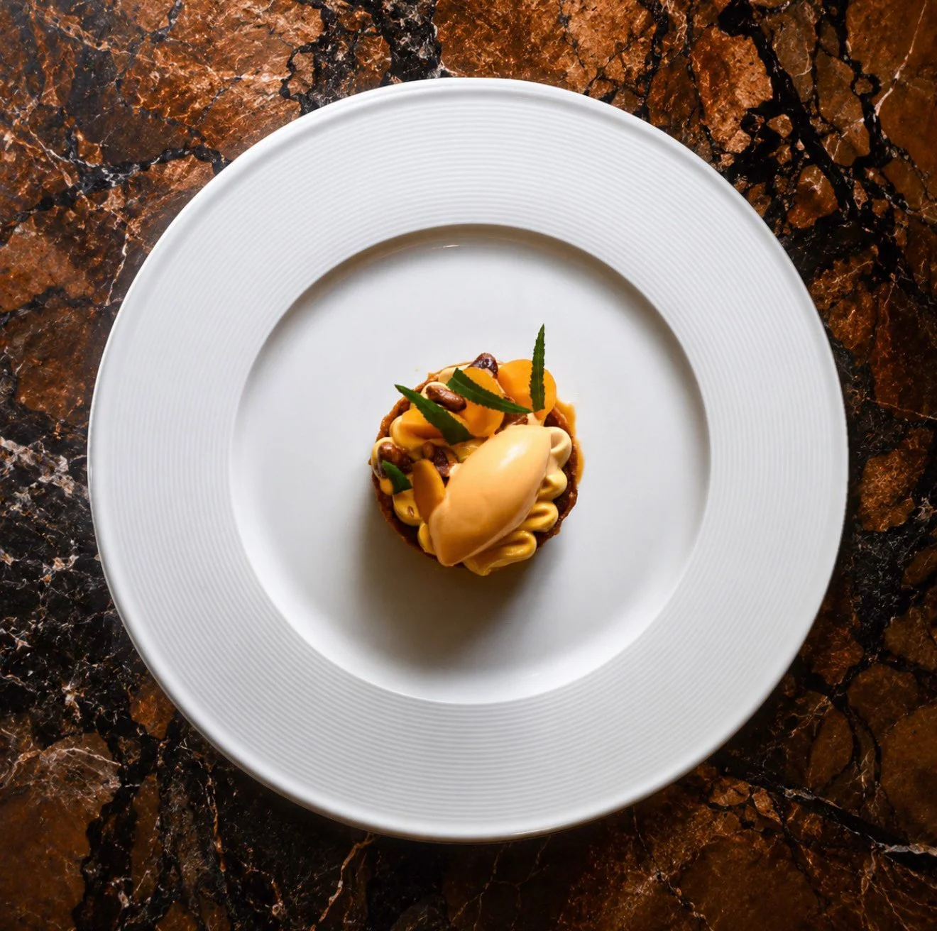 A dessert with yellow and brown toppings, garnished with small green leaves, served on a white plate on a brown marble surface.