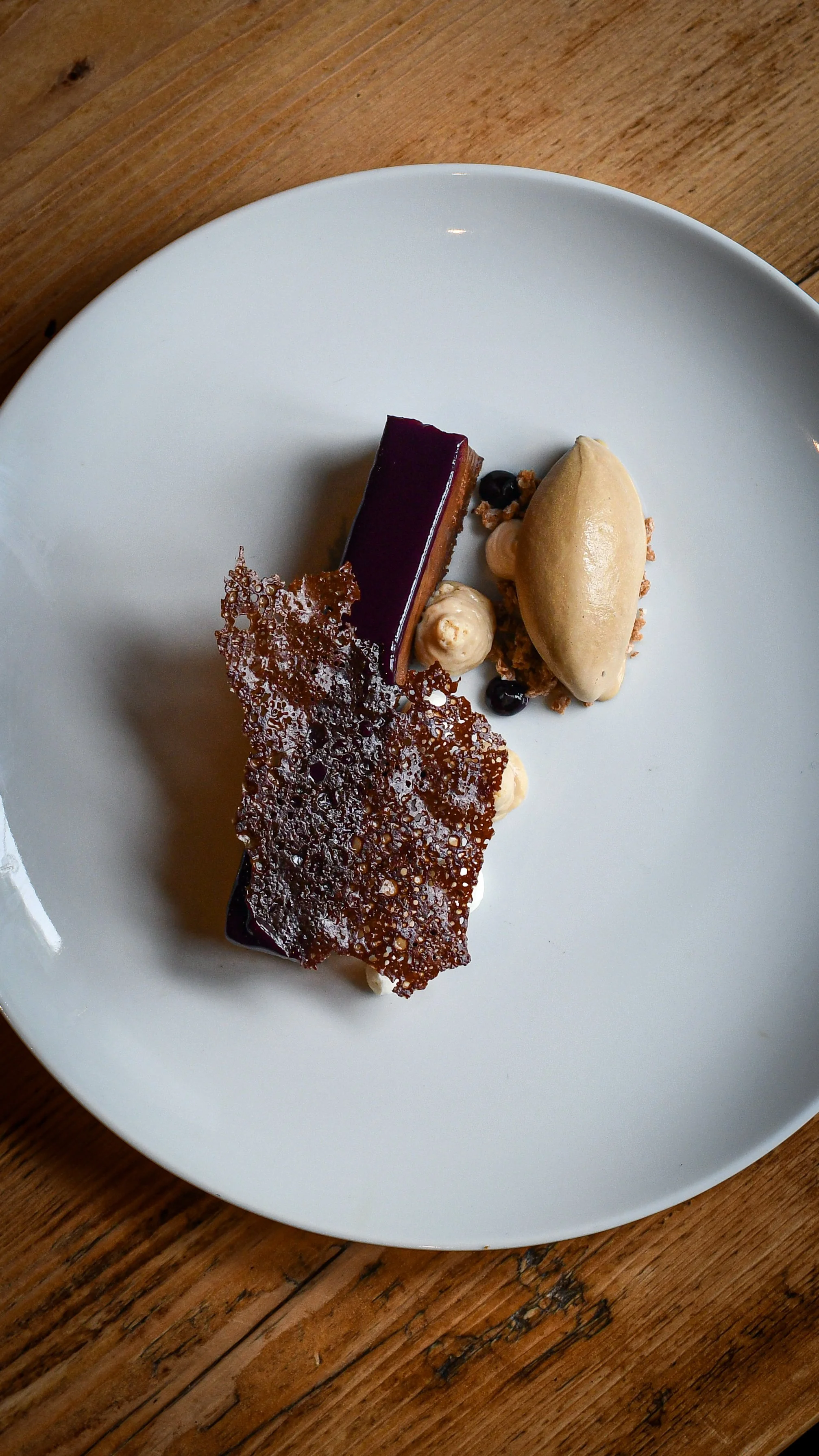A dessert plate with a rectangular chocolate slice, a quenelle of ice cream, caramelized sugar garnish, and small dollops of cream or mousse, on a white plate on a wooden table.