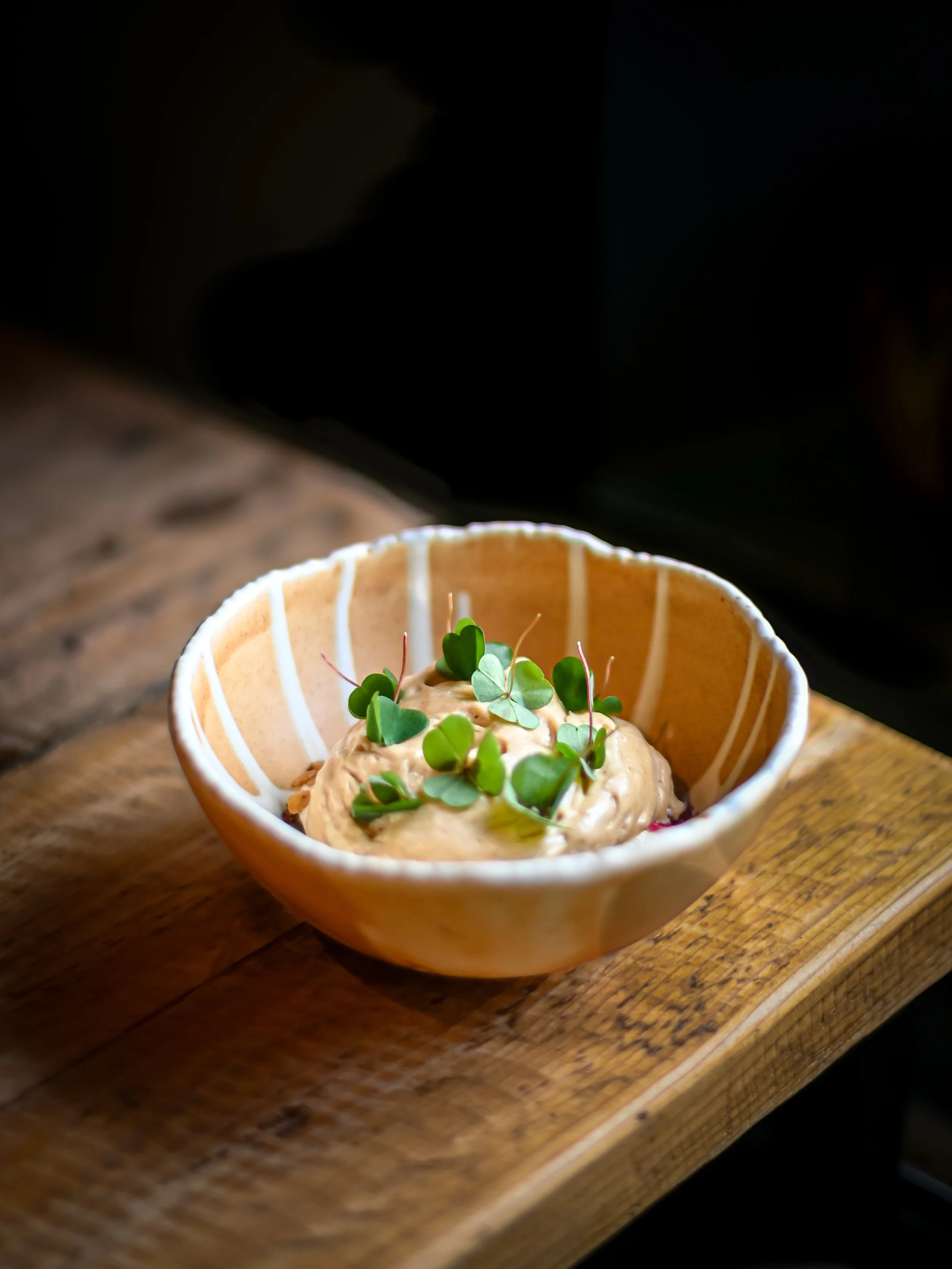 Creamy dip garnished with microgreens in a beige bowl with white stripes, placed on a wooden surface.