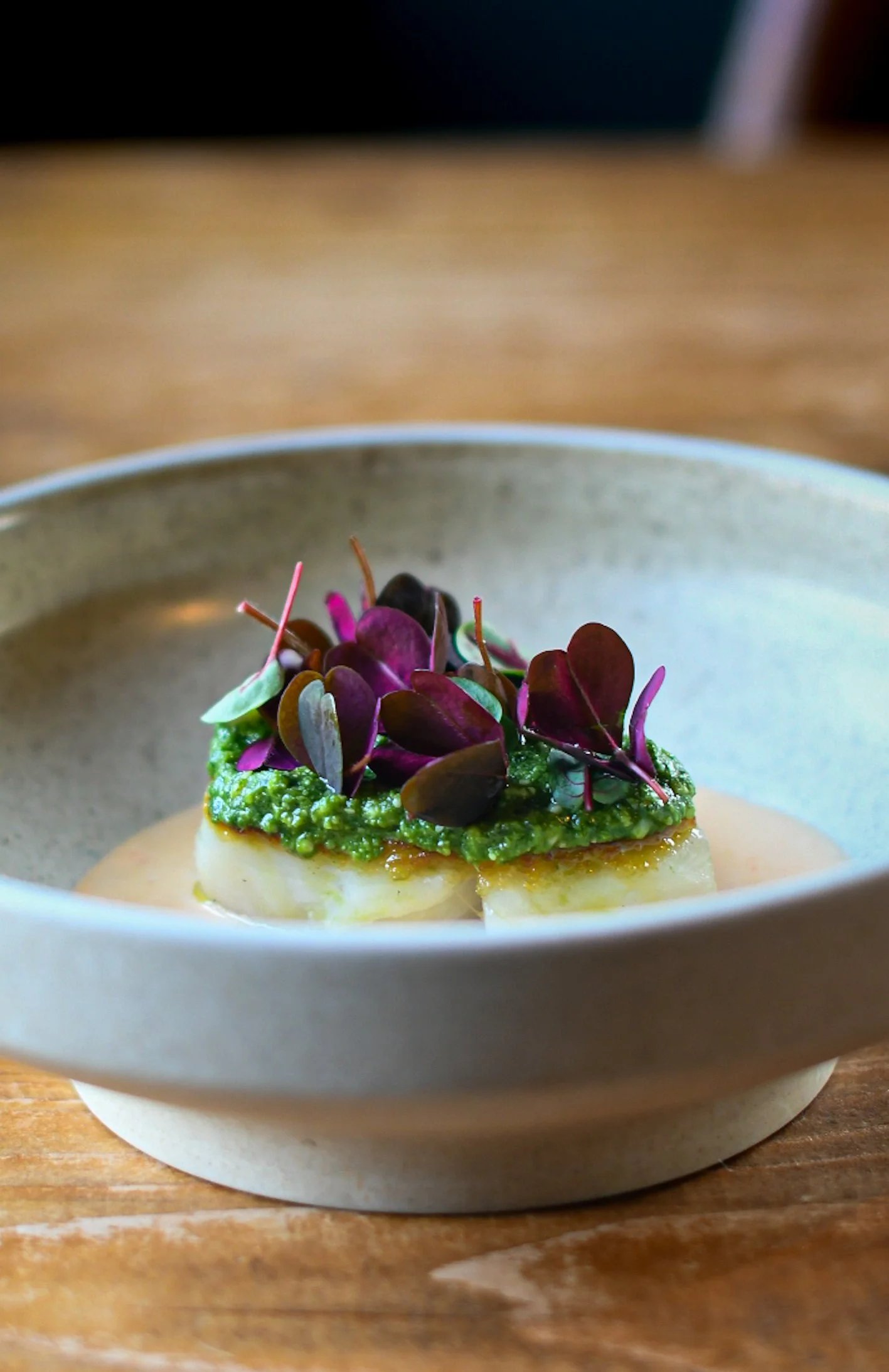 Close-up of an appetizer in a ceramic bowl, featuring a slice of cheese topped with green sauce and purple microgreens, on a wooden surface.