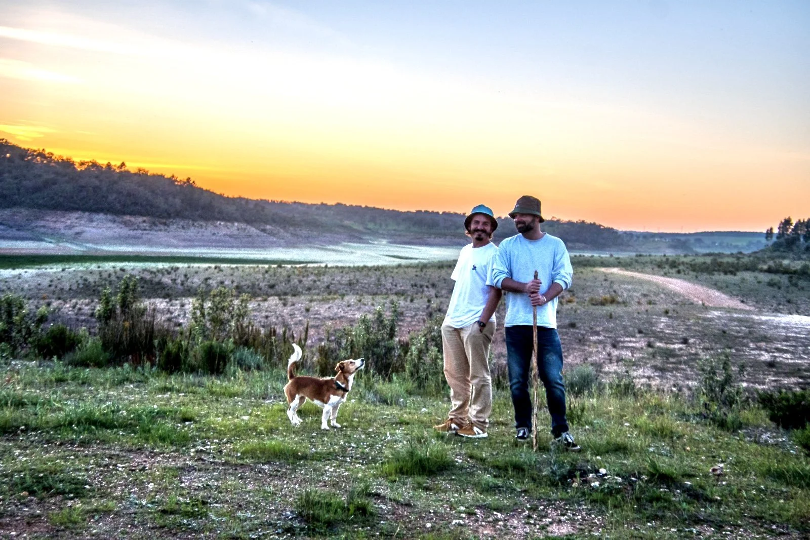Two men with a dog standing outdoors at sunset in a natural landscape with shrubs and rolling hills.