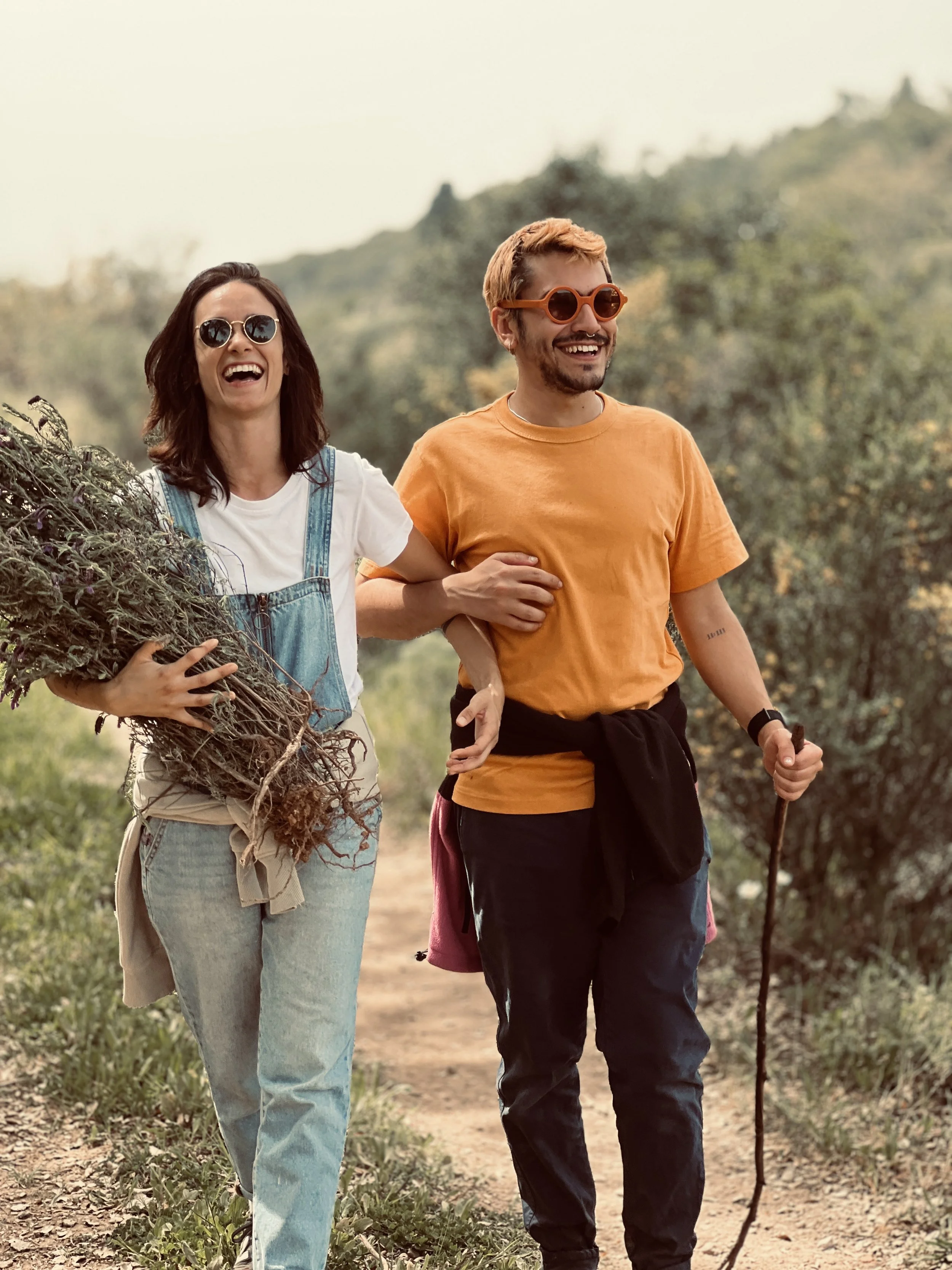 A smiling woman carrying a bundle of plants and a smiling man with a walking stick walking outdoors on a trail in a scenic, hilly area.