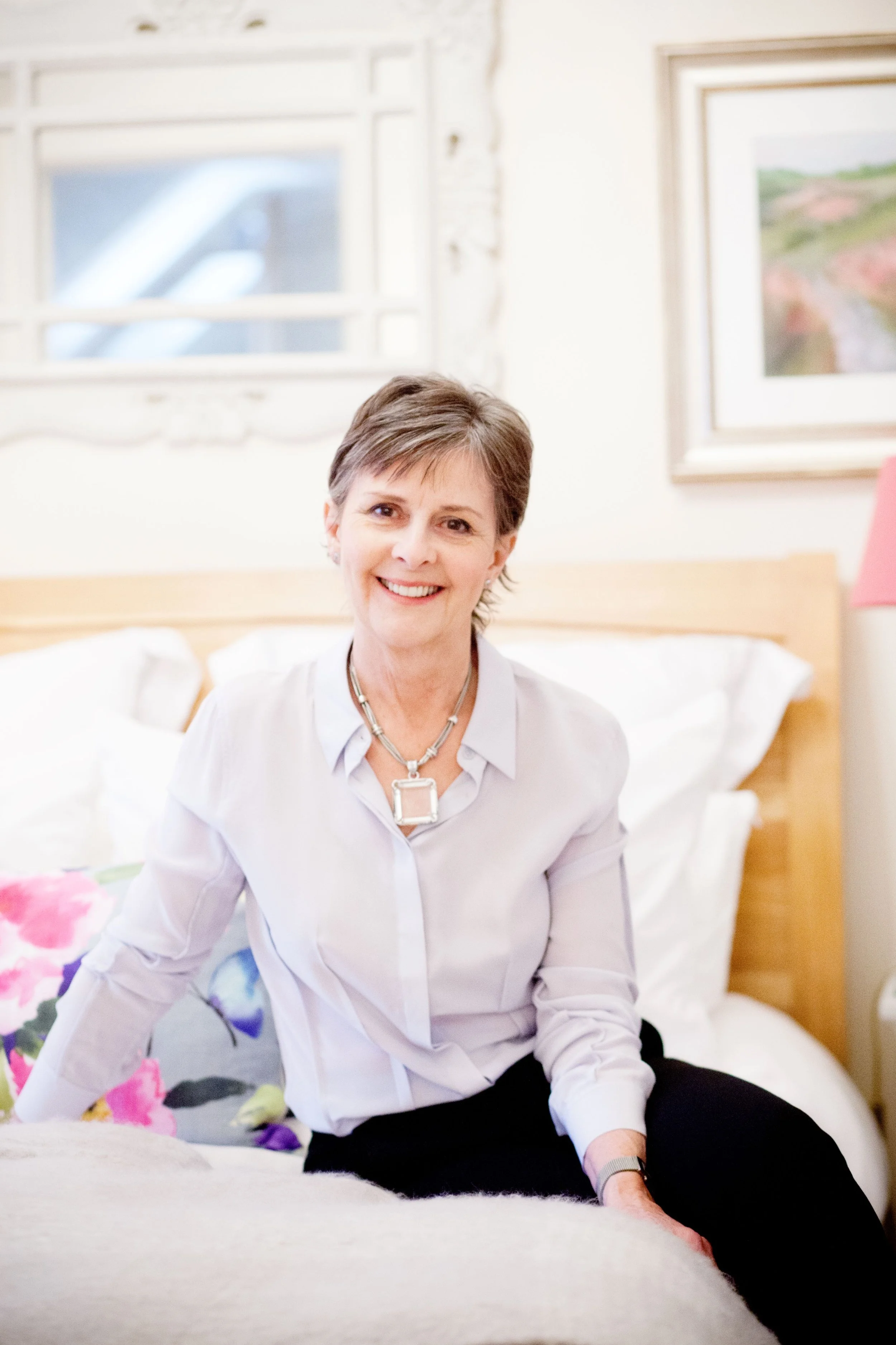 An older lady smiling sat on a bed beautifully dressed.