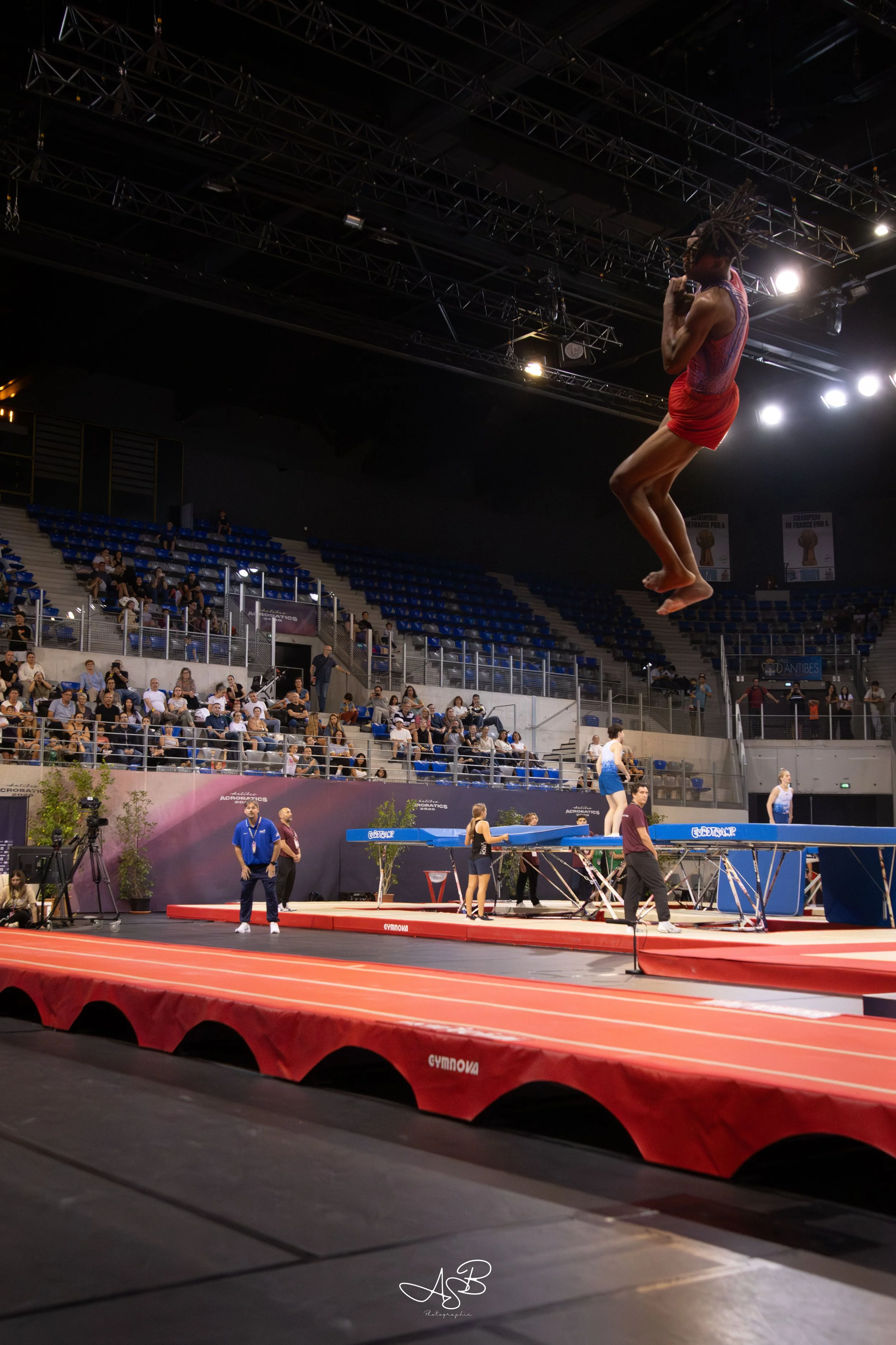 Une femme en saut en hauteur lors d'une compétition de gymnastique dans une grande salle, avec un public assis dans les gradins, et des entraîneurs ou officiels en arrière-plan