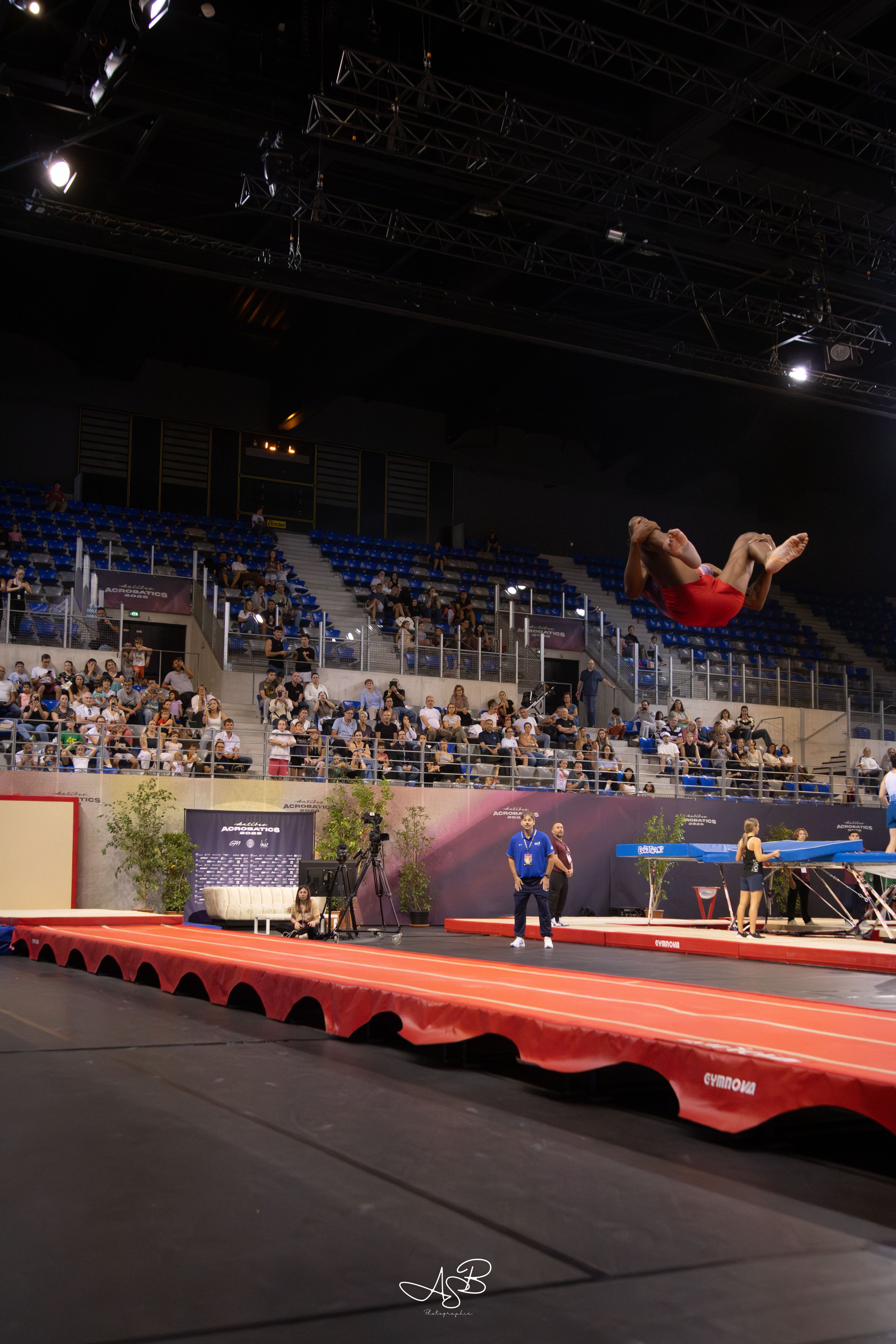 Une gymnaste exécutant une figure dans un gymnase pendant une compétition de gymnastique artistique, avec un public dans les gradins et des appareils de gymnastique en arrière-plan.