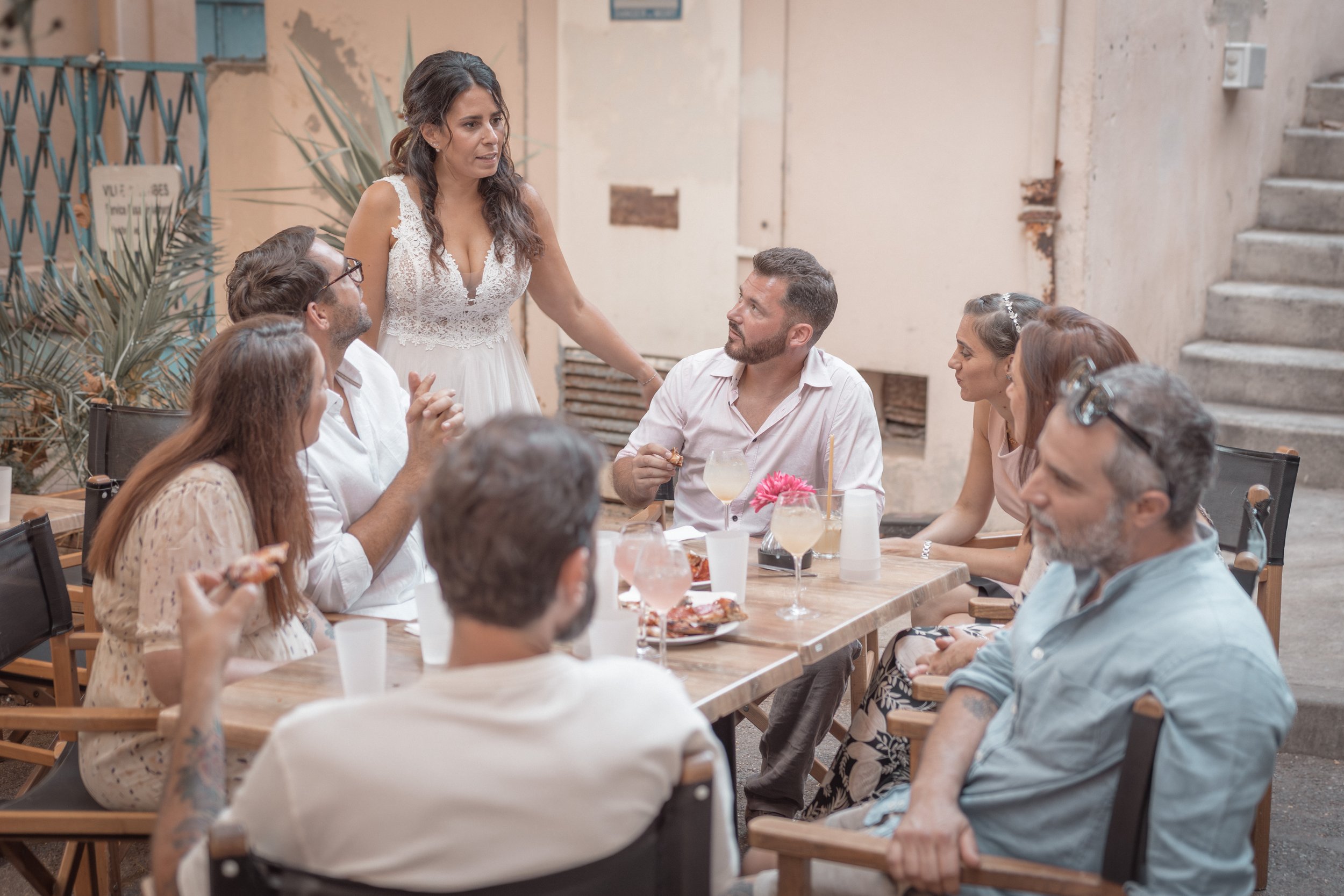 Groupe de personnes assises autour d'une table lors d'une réunion ou célébration en extérieur, avec une femme debout parlant aux autres.