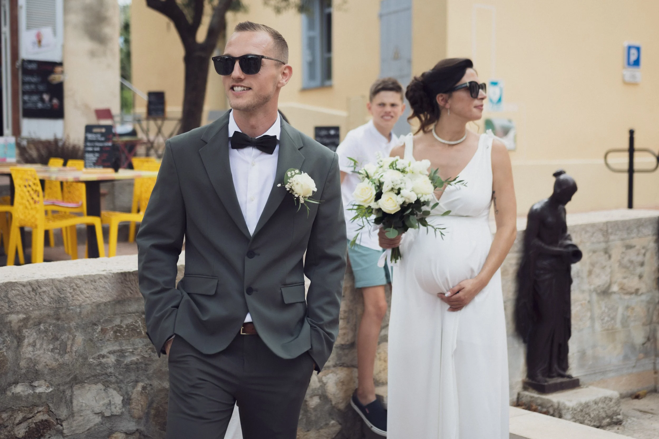 Un groupe de personnes habillées pour un mariage, comprenant un homme en costume gris avec nœud papillon et une femme en robe blanche avec bouquet, rencontrent ou assistent à une cérémonie à l'extérieur.