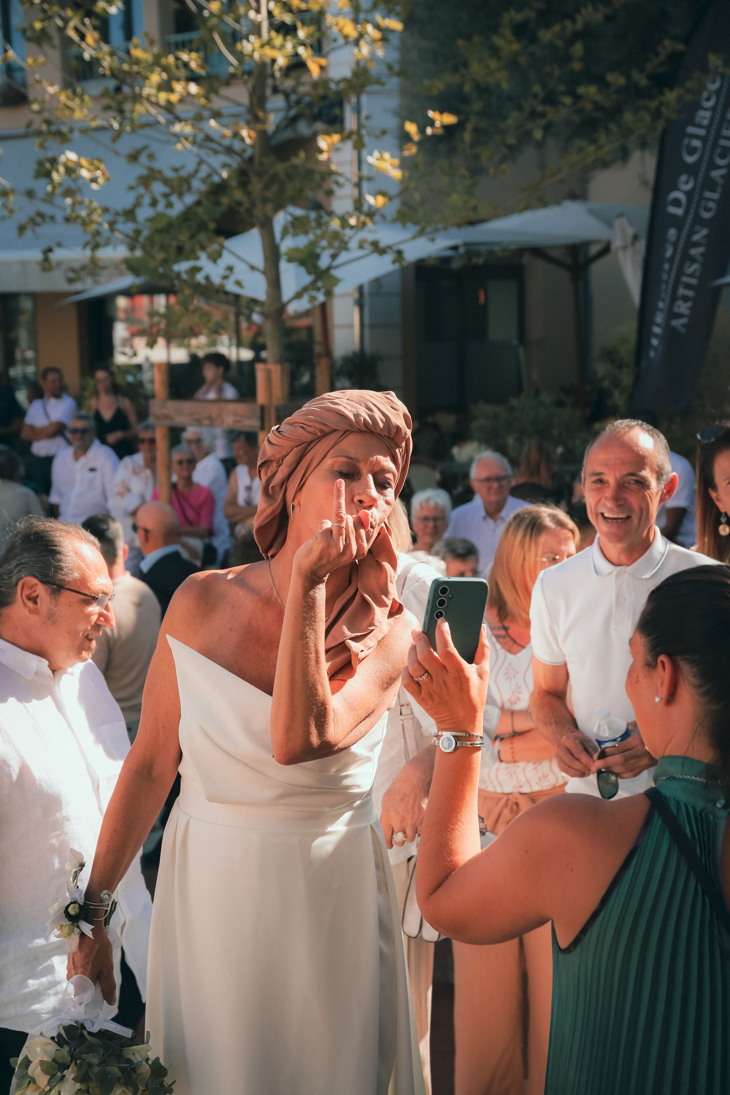 Une femme portant une robe blanche et un turban beige tient un téléphone mobile, levant son doigt en signe de défi ou de signature, entourée d’un groupe de personnes souriantes lors d’un événement en plein air, éclairé par la lumière du soleil.