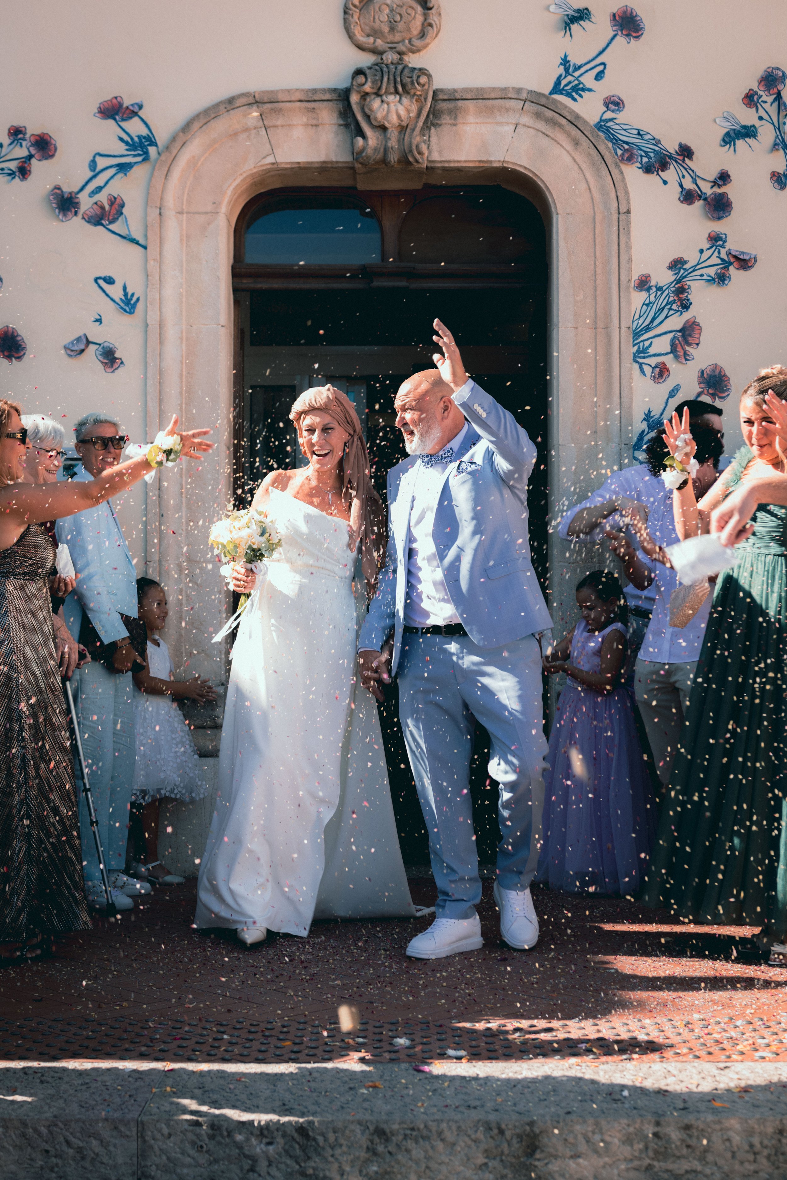 Un mariage avec un couple de mariés, entourés d'invités, lançant des confettis devant une entrée de bâtiment ornée de fleurs murales.