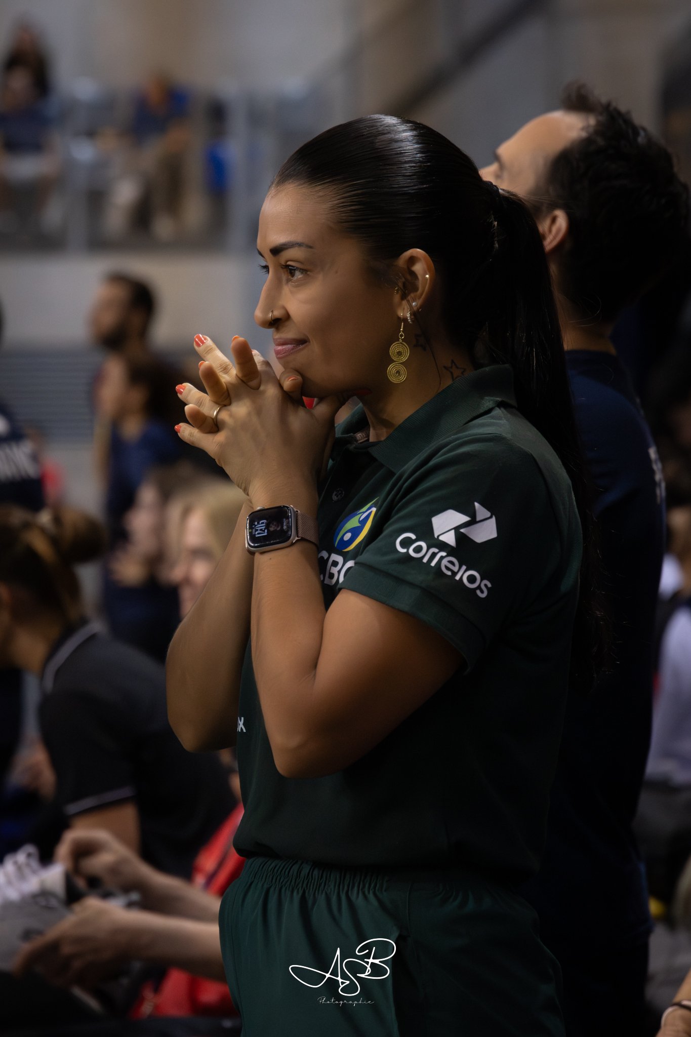 Une femme regardant fixement un événement sportif, avec un sourire, portant une chemise verte avec des logos, une montre intelligente, et des boucles d'oreilles dorées, dans une salle remplie de spectateurs.
