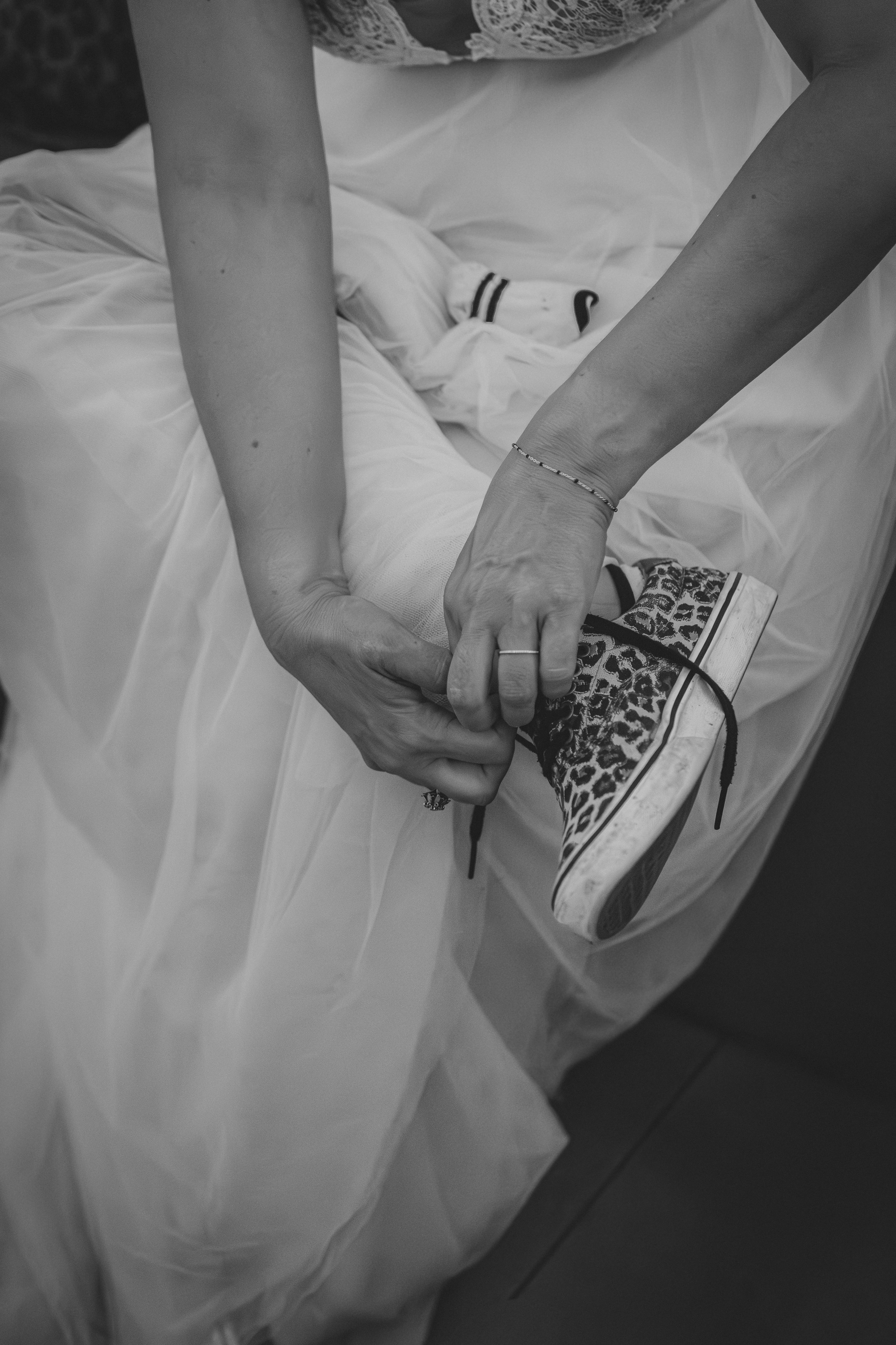 Une personne aide une autre à attacher ses chaussures, avec une mise en scène intime sur un lit. La personne aidante porte un bracelet et une bague, et celle qui est assistée a une jambe en plâtre.