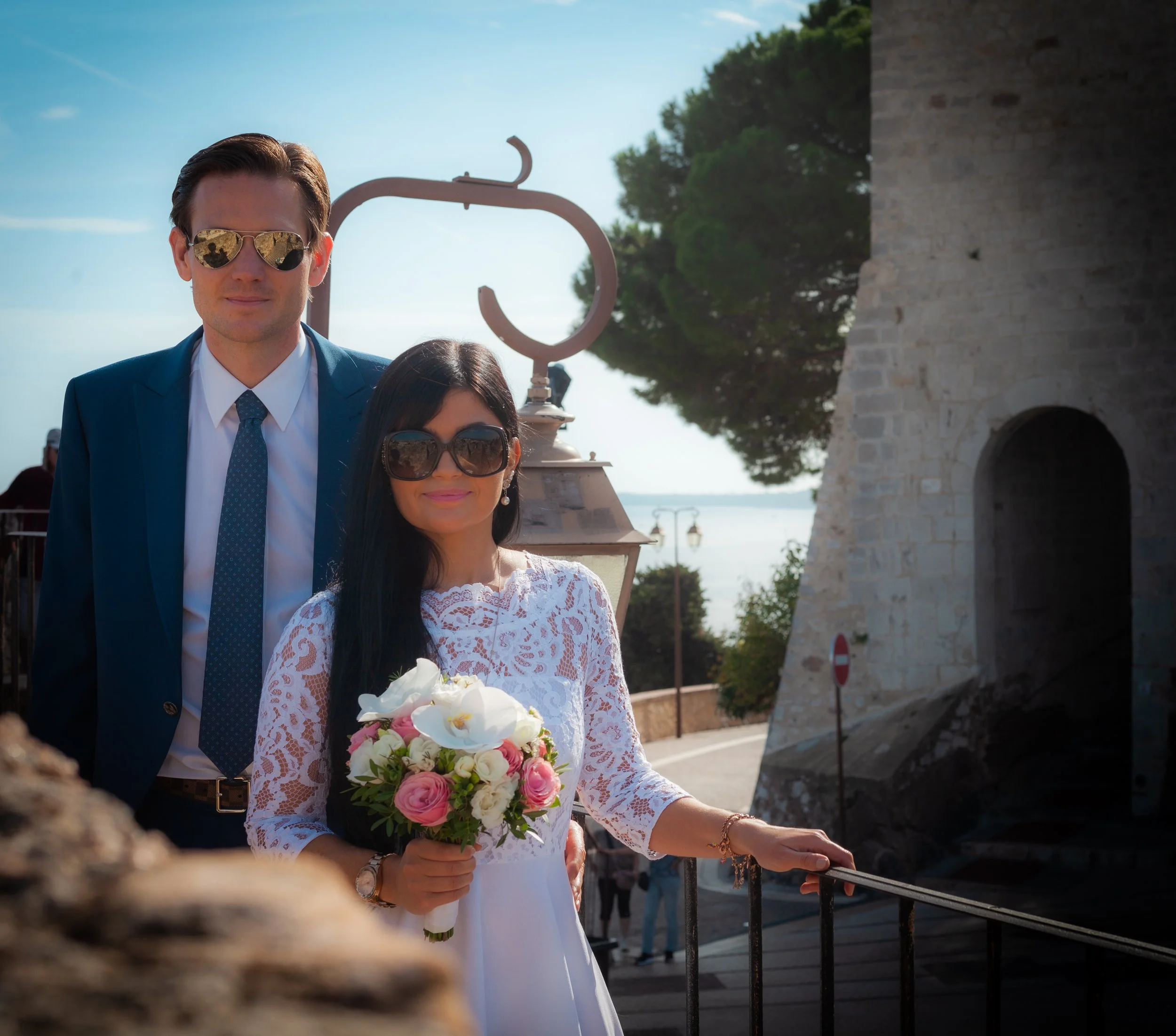 Un couple habillé pour un mariage, la femme tenant un bouquet de fleurs, la femme portant des lunettes de soleil et un sourire, la scène en plein air avec des arbres et une mer en arrière-plan.