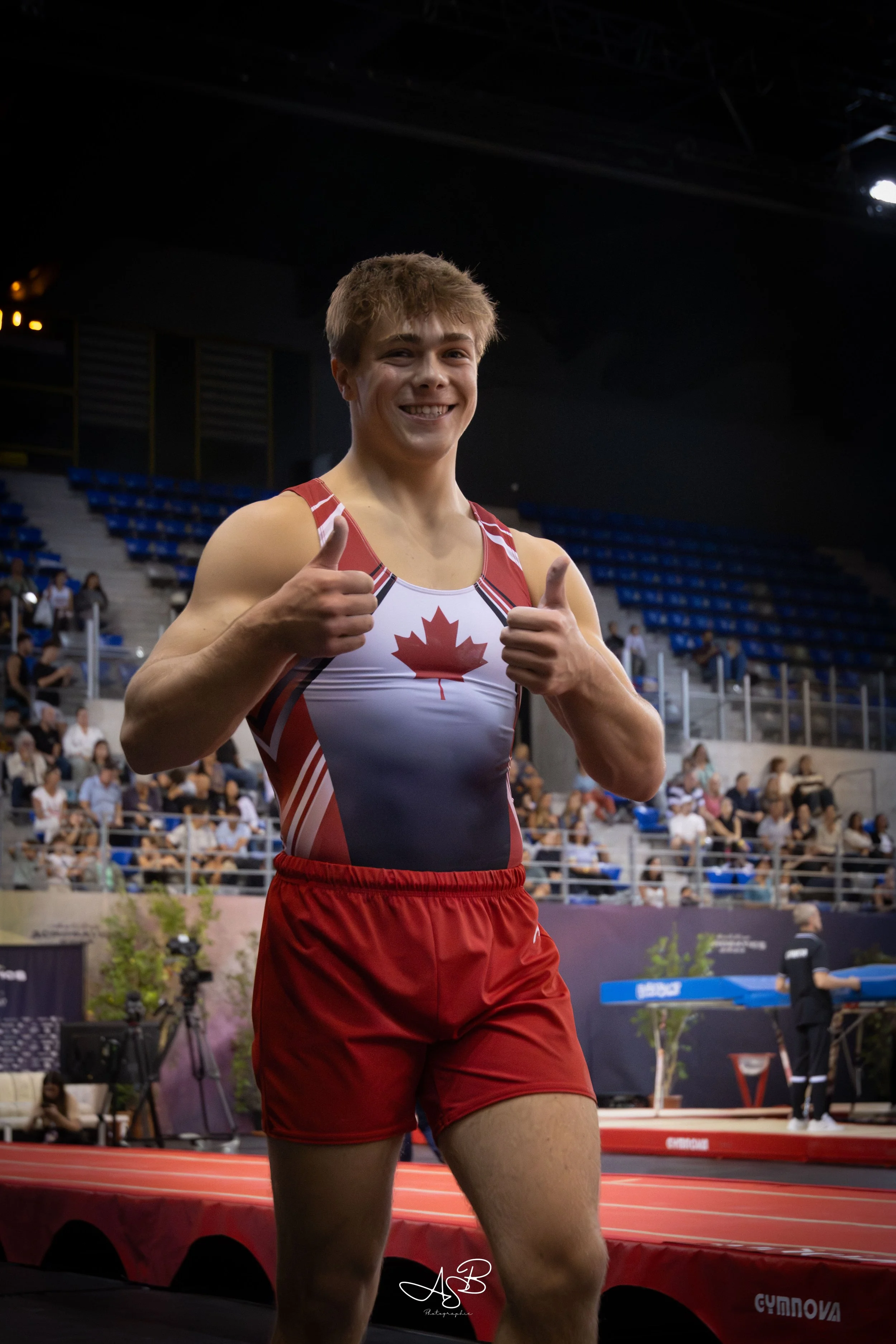 Jeune athlète masculin canadien en uniforme rouge et blanc, souriant et faisant un signe de pouce en l'air, lors d'une compétition d'athlétisme en intérieur, avec des spectateurs en arrière-plan.
