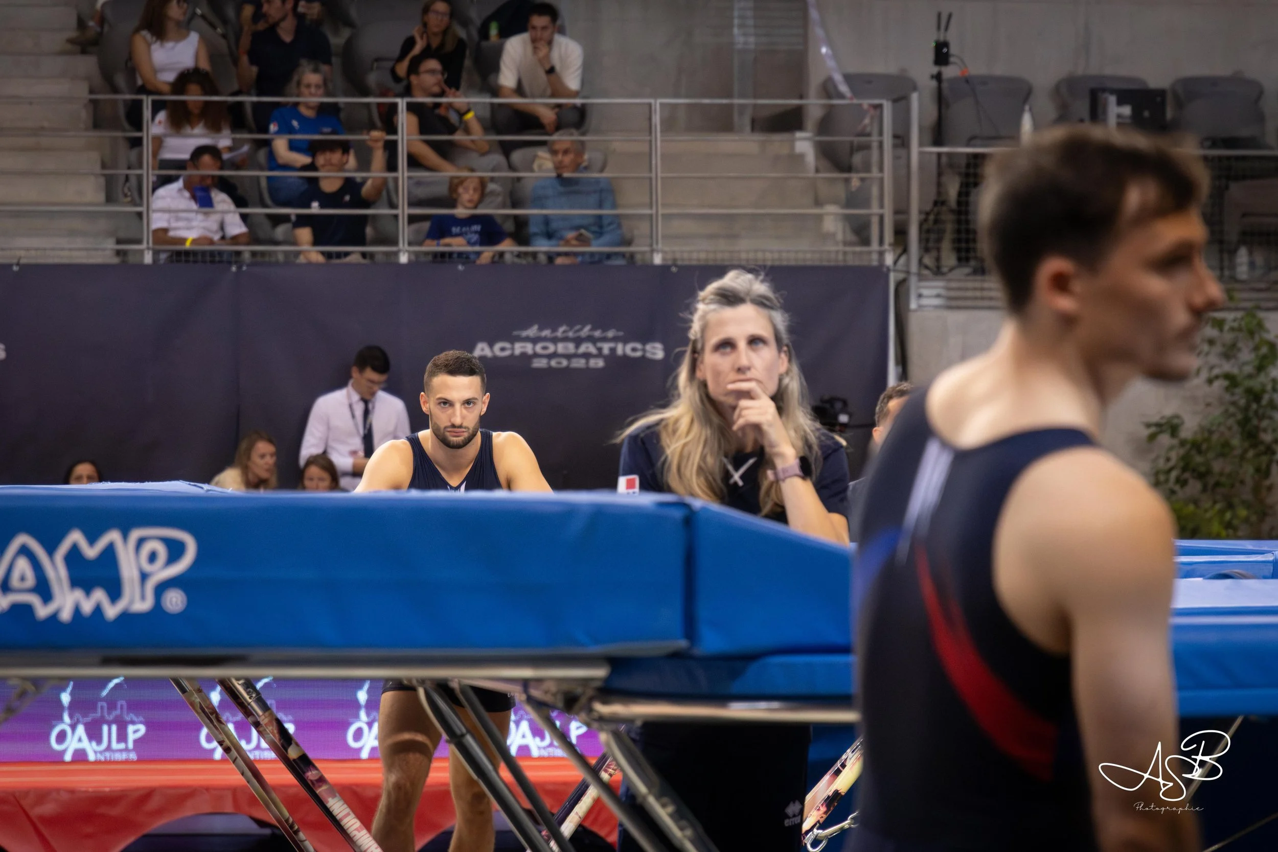 Pendant une compétition de saut en trampoline, un homme en tenue de sport se tient à côté d'une table avec un regard concentré. Une femme assise derrière lui, également en tenue de sport, semble réfléchir ou analyser la situation. En arrière-plan, d'