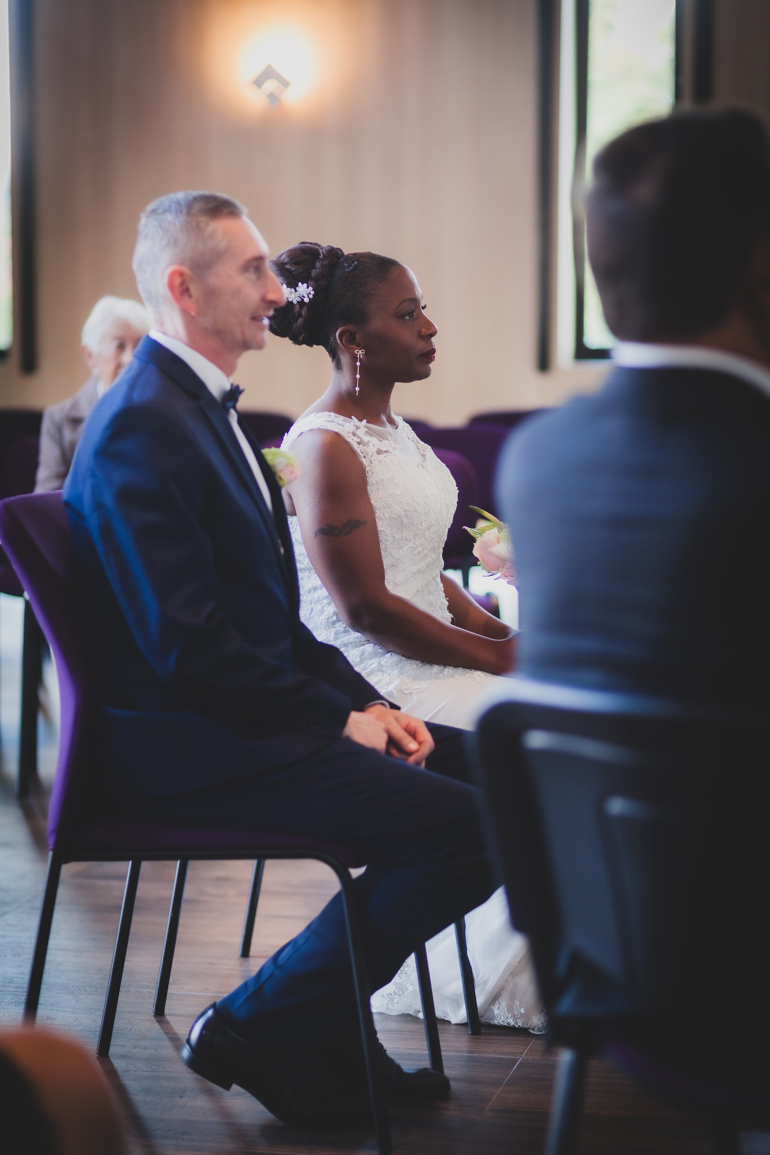 Un couple assis lors d'une cérémonie de mariage, la femme en robe de mariée blanche, l'homme en costume noir, dans une salle lumineuse avec des fenêtres