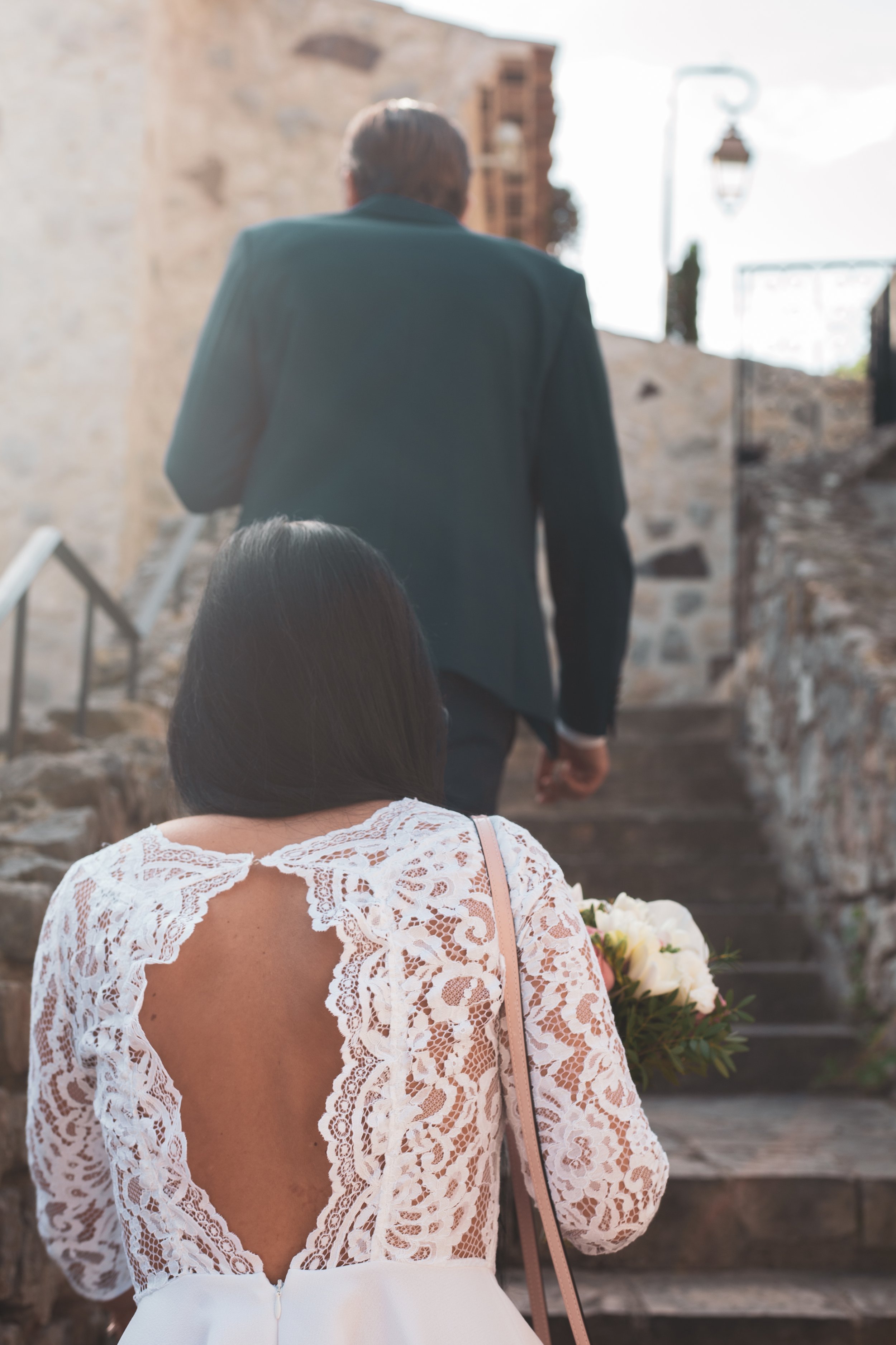 Une femme en robe blanche avec un dos en dentelle regarde un homme en costume noir monter des escaliers en pierre dans un lieu ancien.