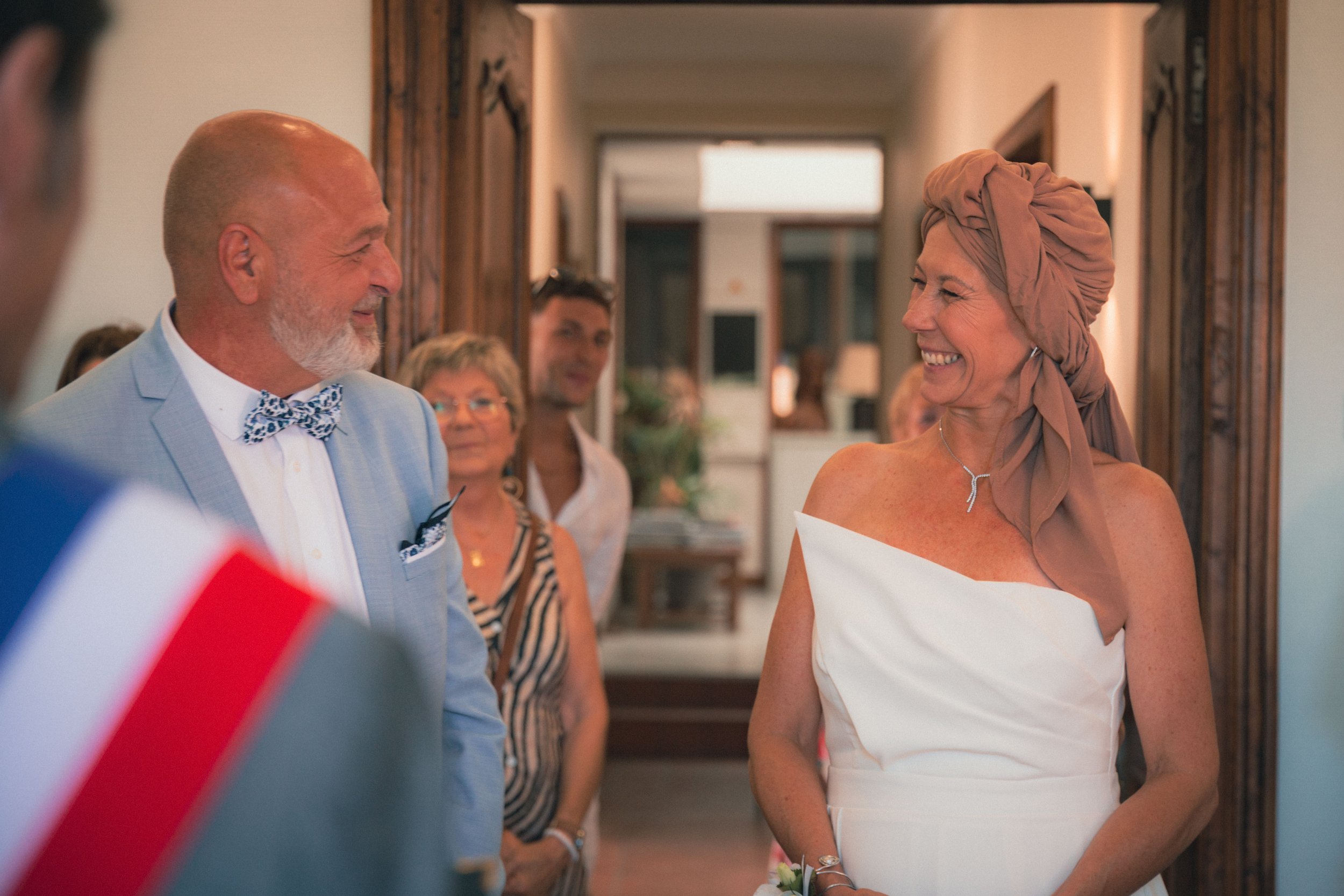 Une femme en robe blanche sourit à un homme en costume bleu pastel avec un nœud papillon à motifs, lors d'un mariage ou d'une célébration, entourés de plusieurs personnes dans une maison.