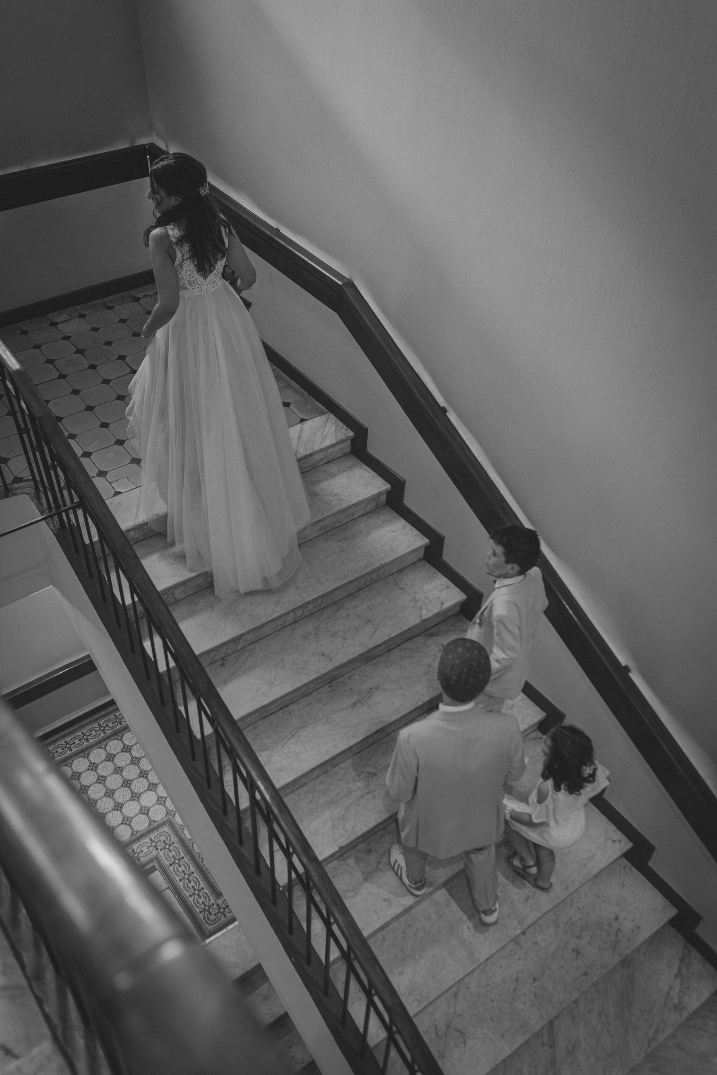 Une femme en robe de mariée descend un escalier, entourée de plusieurs personnes.