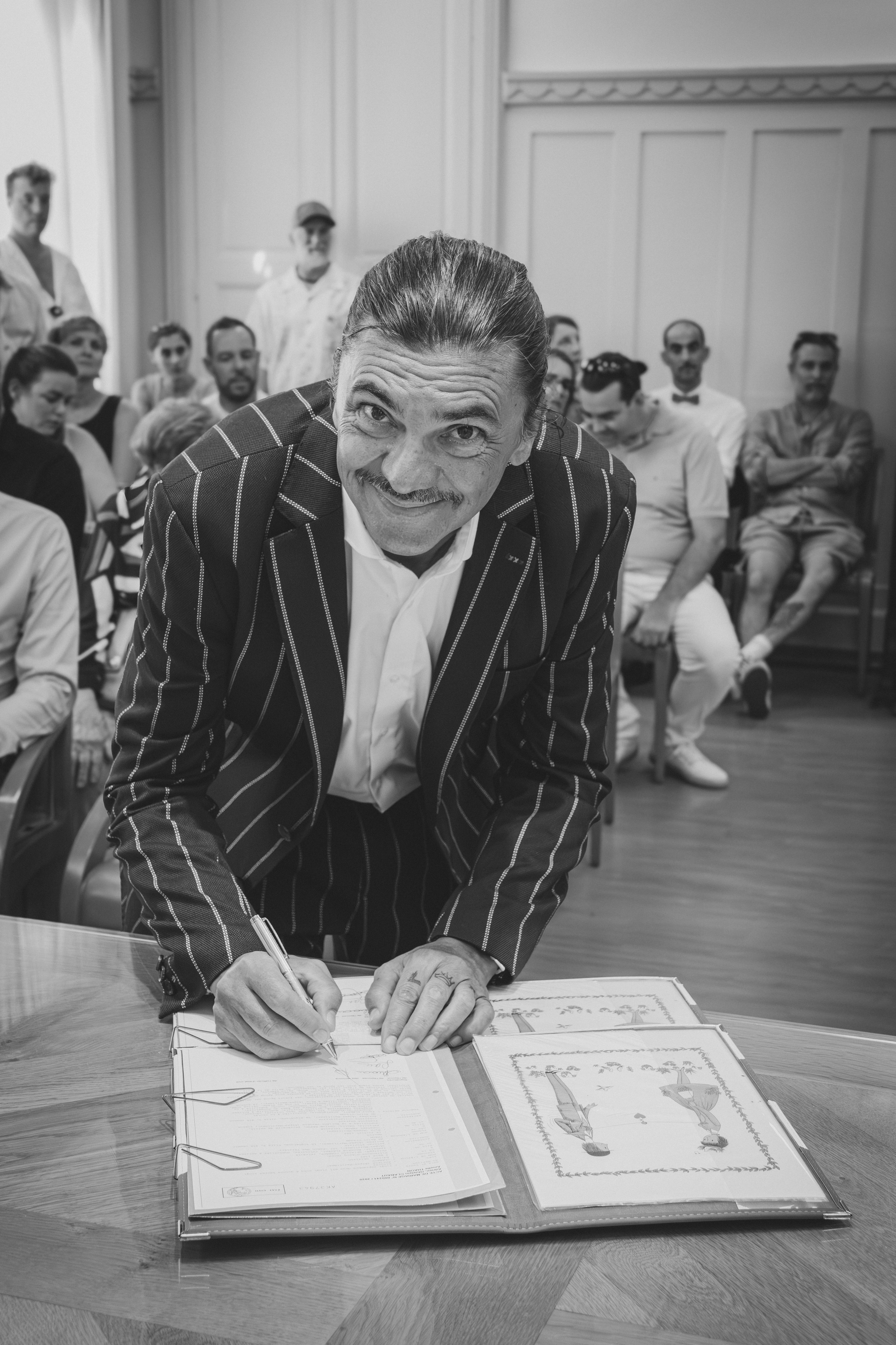 Un homme en costume à rayures écrit dans un livre à une table, entouré par une audience assise dans une pièce élégante.