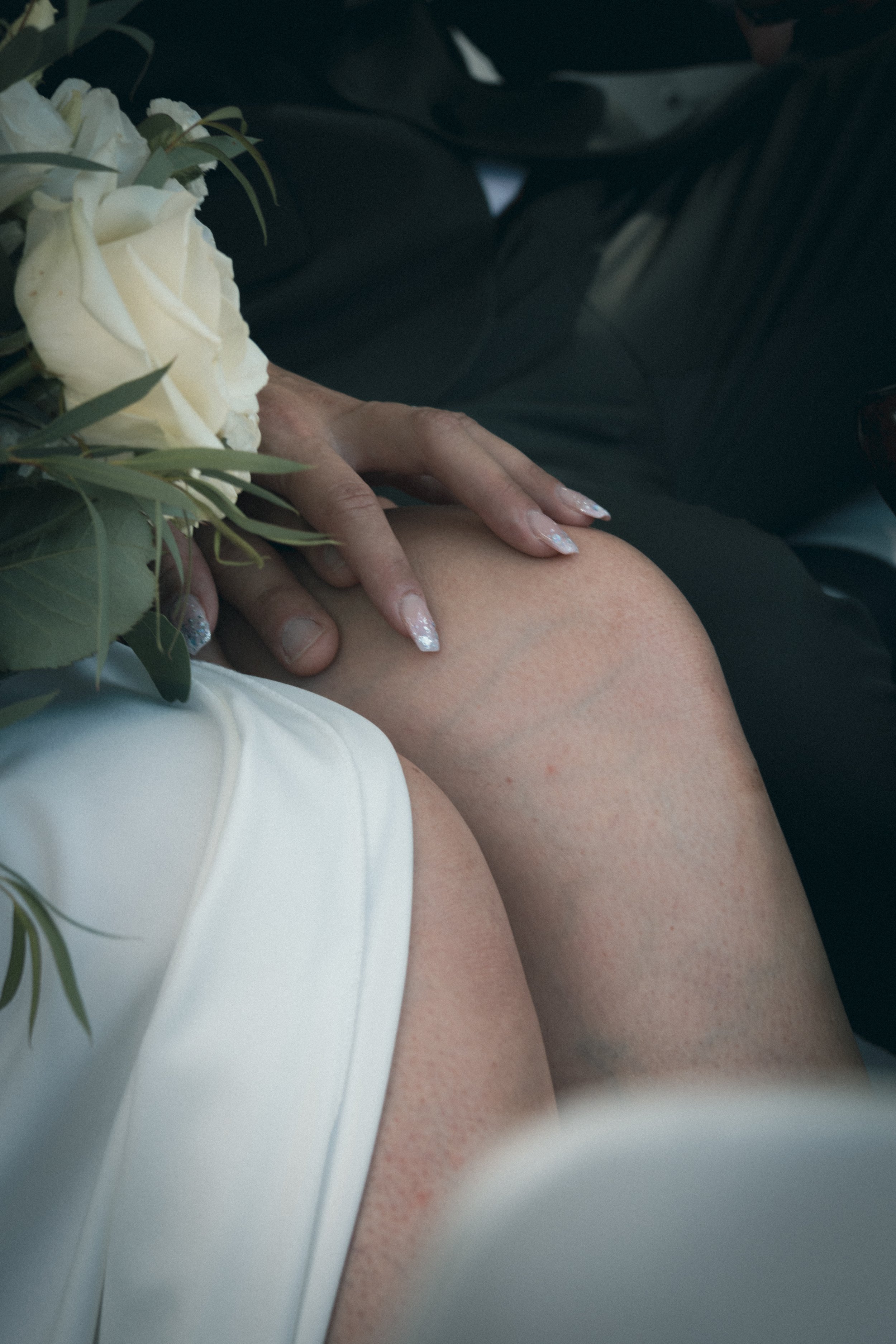 Détail d'une main tenant une jambe, avec un bouquet de fleurs blanches, probablement une mariée, dans une ambiance de mariage.