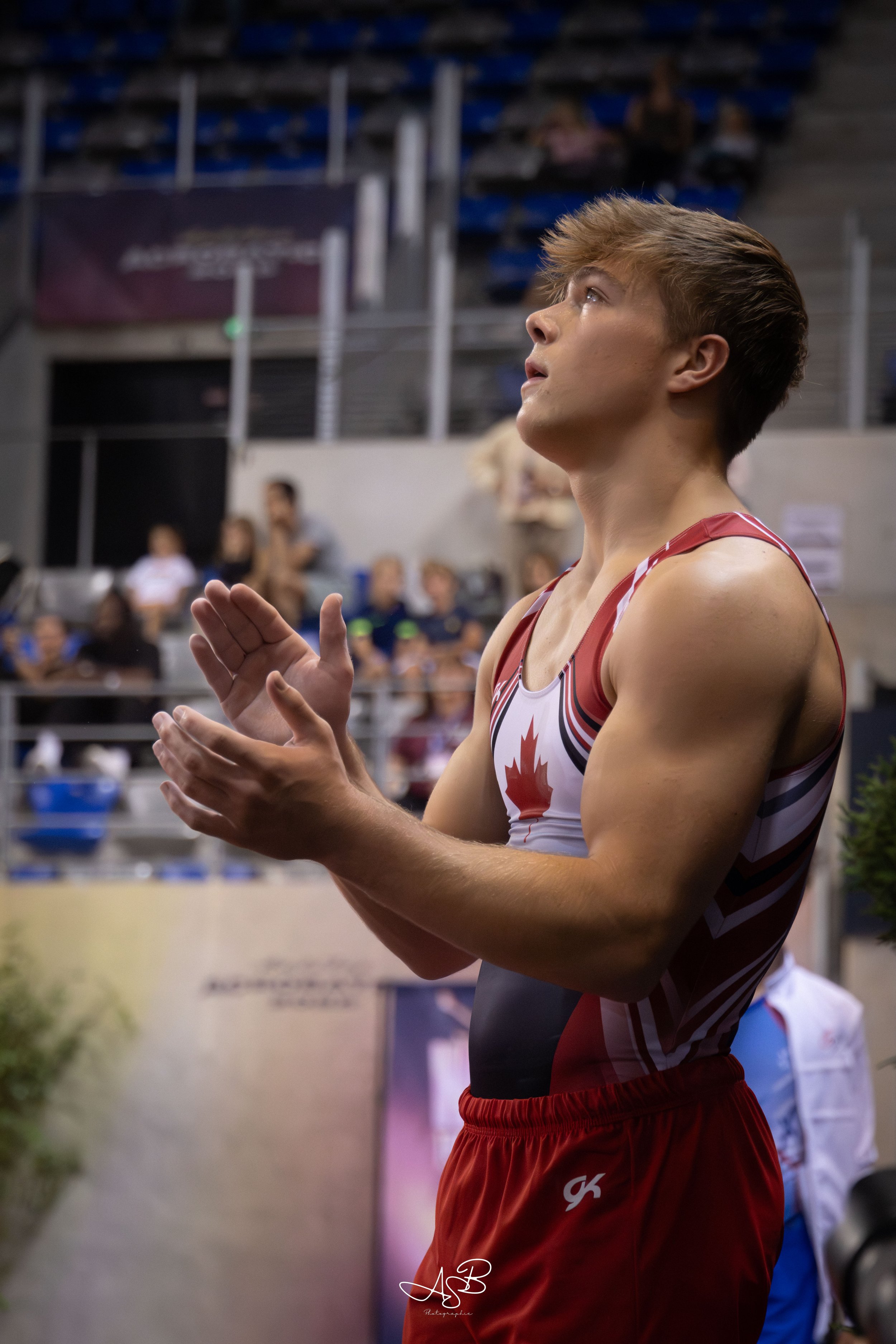 Jeune athlète masculain en tenue sportive avec une feuille d'érable, en train de applaudir lors d'une compétition en intérieur, avec des spectateurs en arrière-plan.