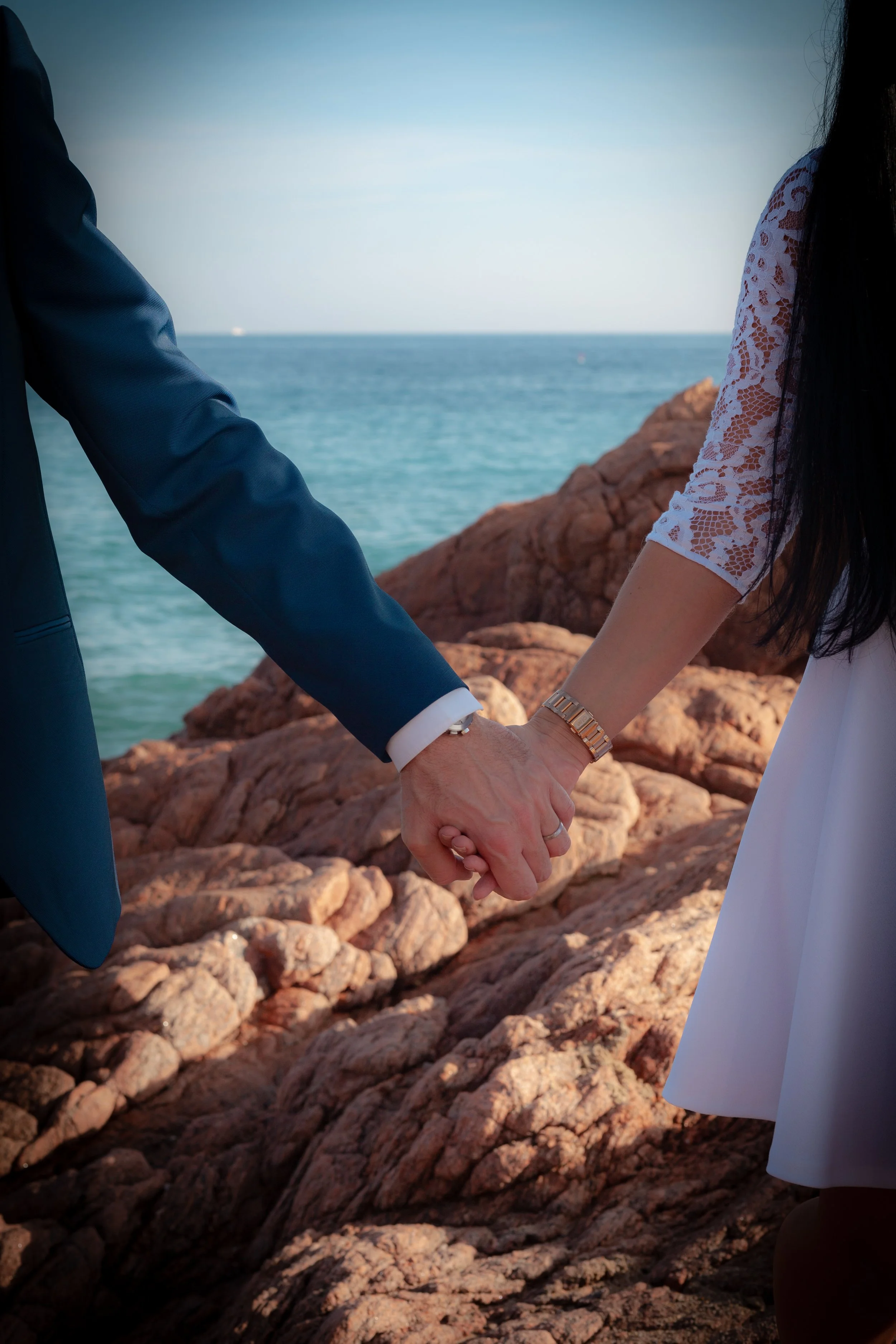 Deux personnes tenant la main au bord de la mer, vues partiellement, une en costume bleu et l'autre en robe blanche, avec des rochers en arrière-plan.