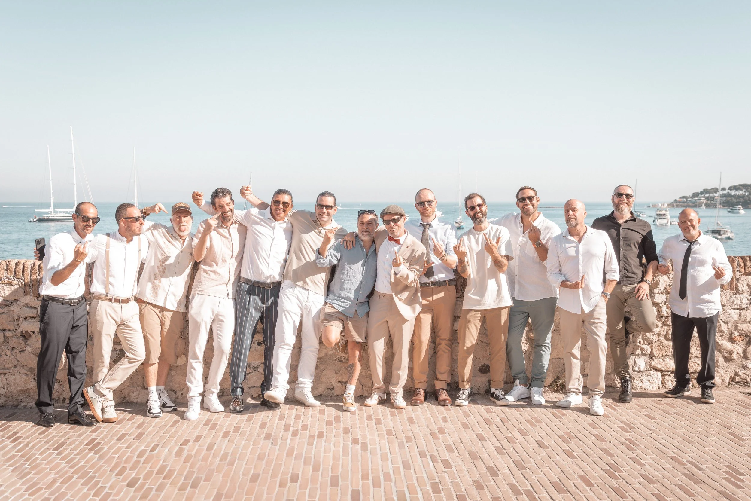 Groupe d'hommes posant en groupe devant un mur en pierre, avec la mer et des bateaux en arrière-plan, lors d'une journée ensoleillée.