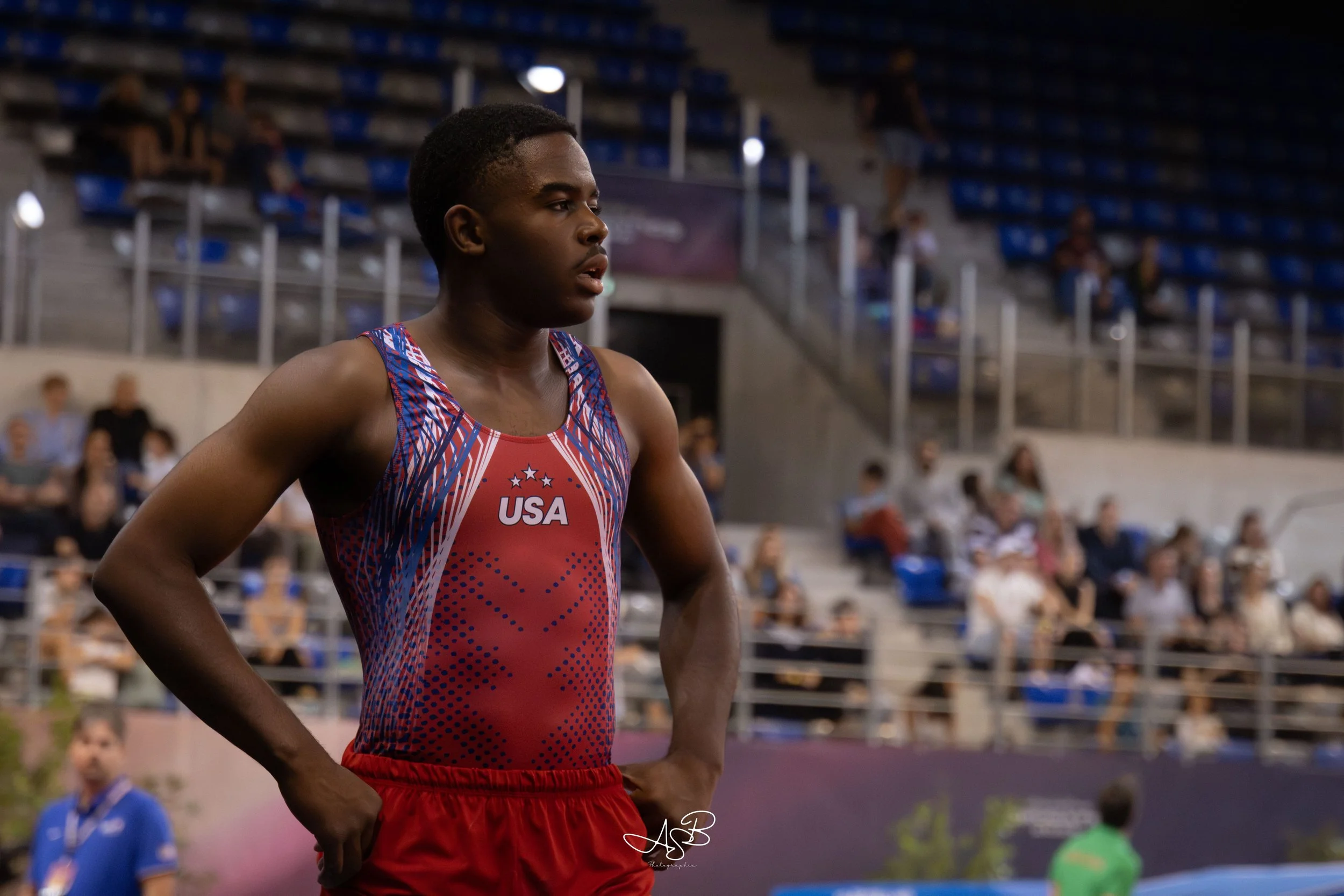 Un athlète américain vêtu d'un maillot rouge et bleu, prêt pour une compétition d'athlétisme, dans un stade intérieur avec des spectateurs en arrière-plan.