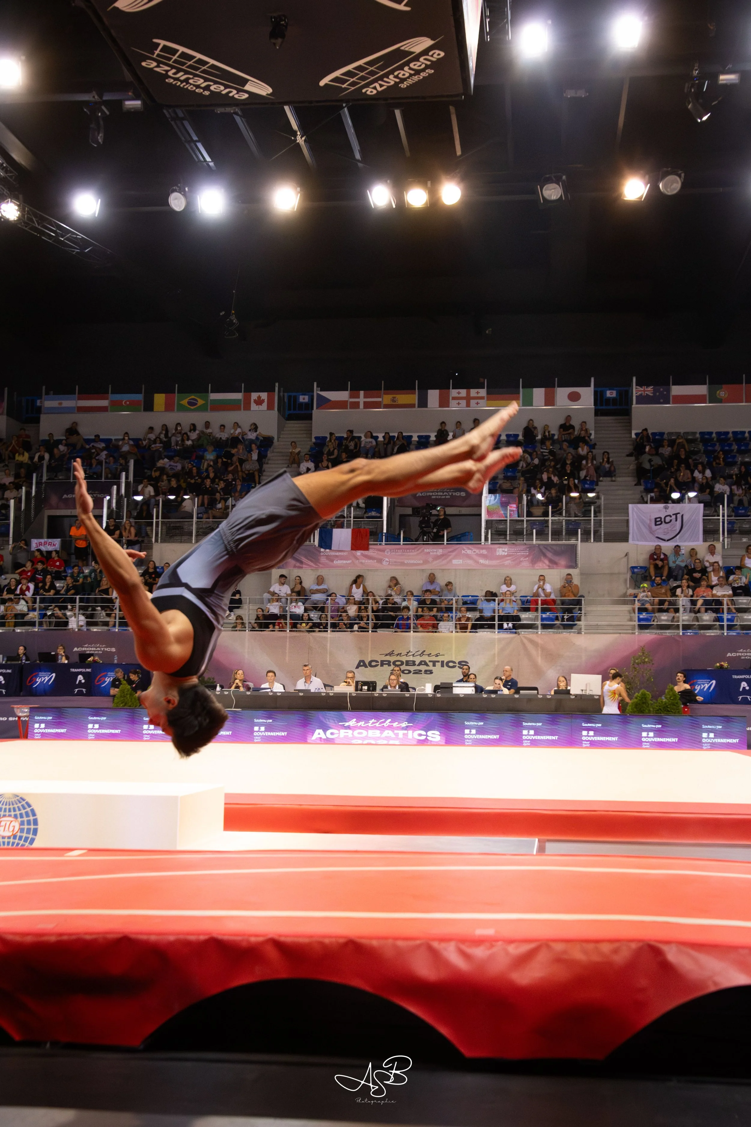 Un gymnaste effectuant un saut acrobatique lors d'une compétition d'acrobatie dans une salle avec un public, drapeaux de différents pays en arrière-plan.