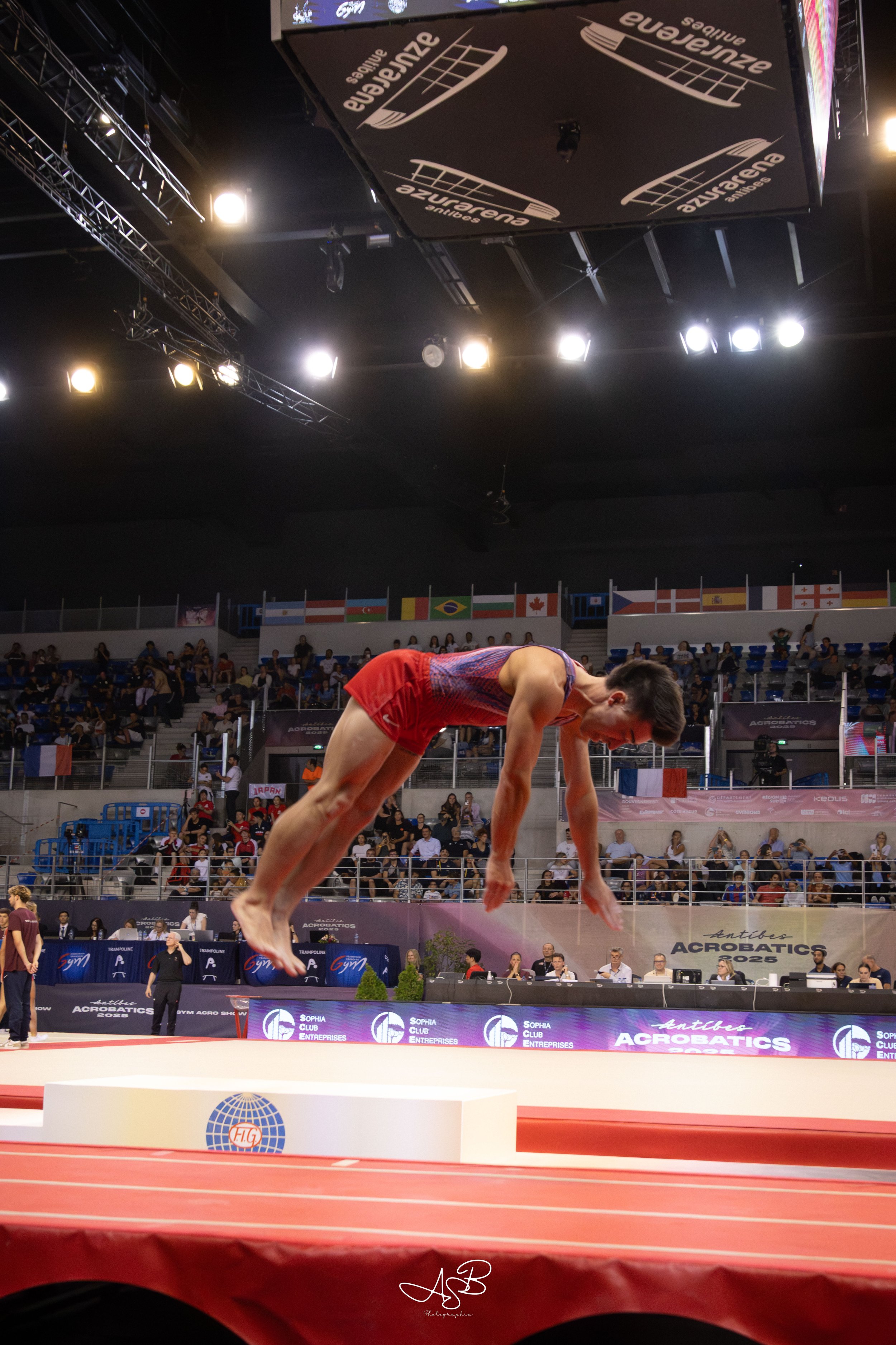 Une gymnaste en plein saut lors d'une compétition d'acrobatie à Antibes, avec un public et des drapeaux internationaux en arrière-plan.