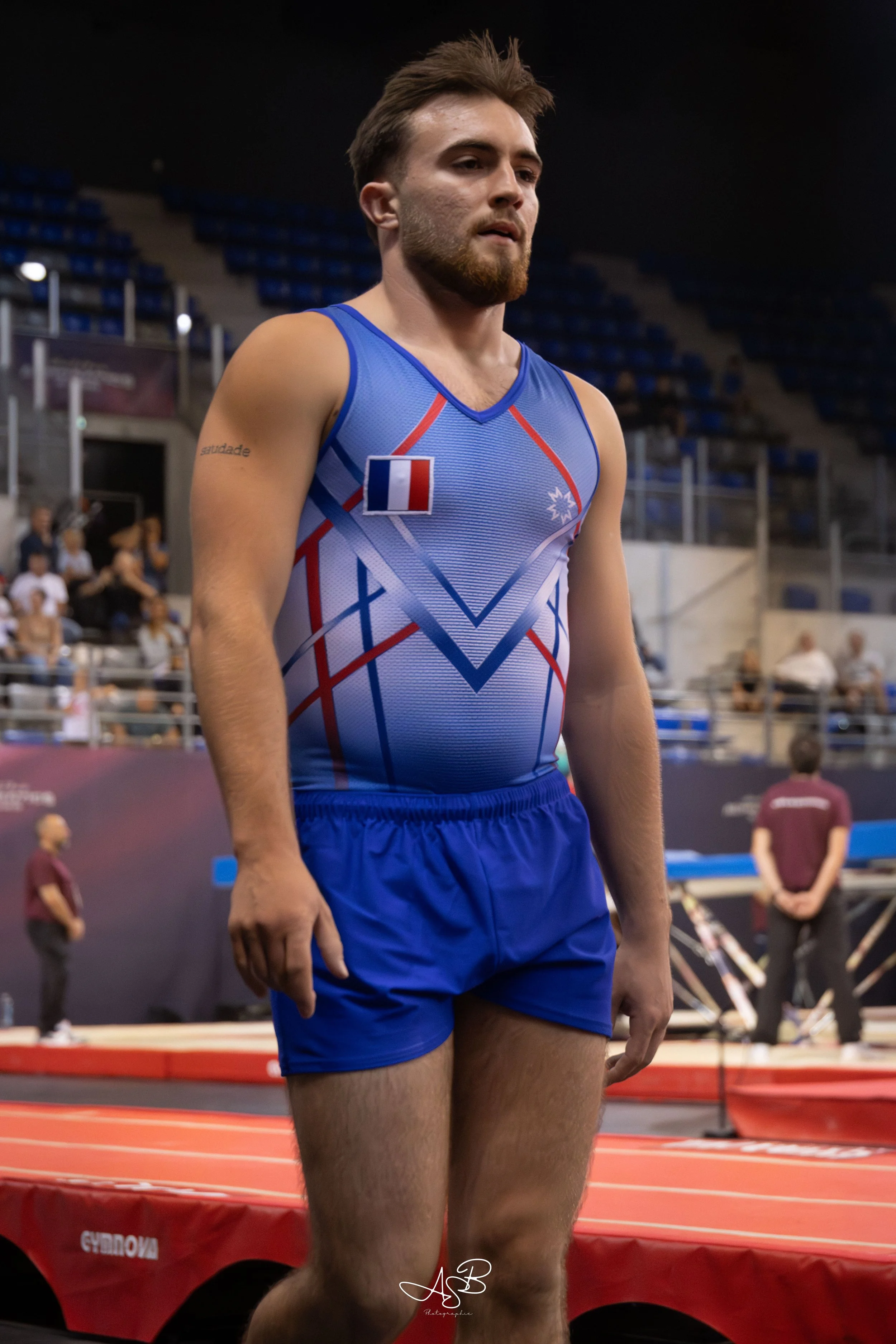 Un athlète masculin en tenue de gymnastique bleue avec un drapeau français sur le torse, dans une salle d'entraînement ou de compétition.