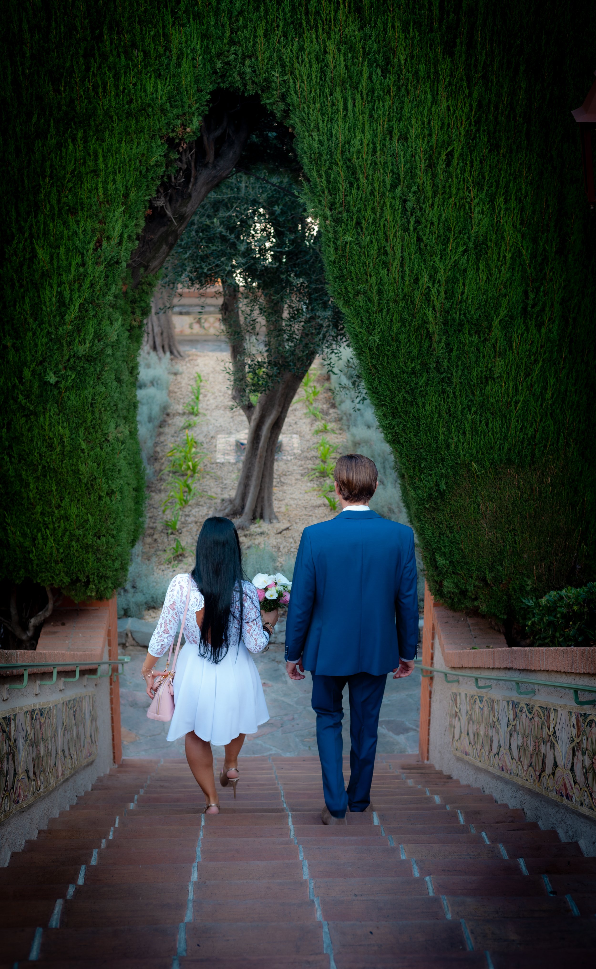 Un couple descend une série de marches en découvrant un jardin avec un arbre et des haies bien taillées.