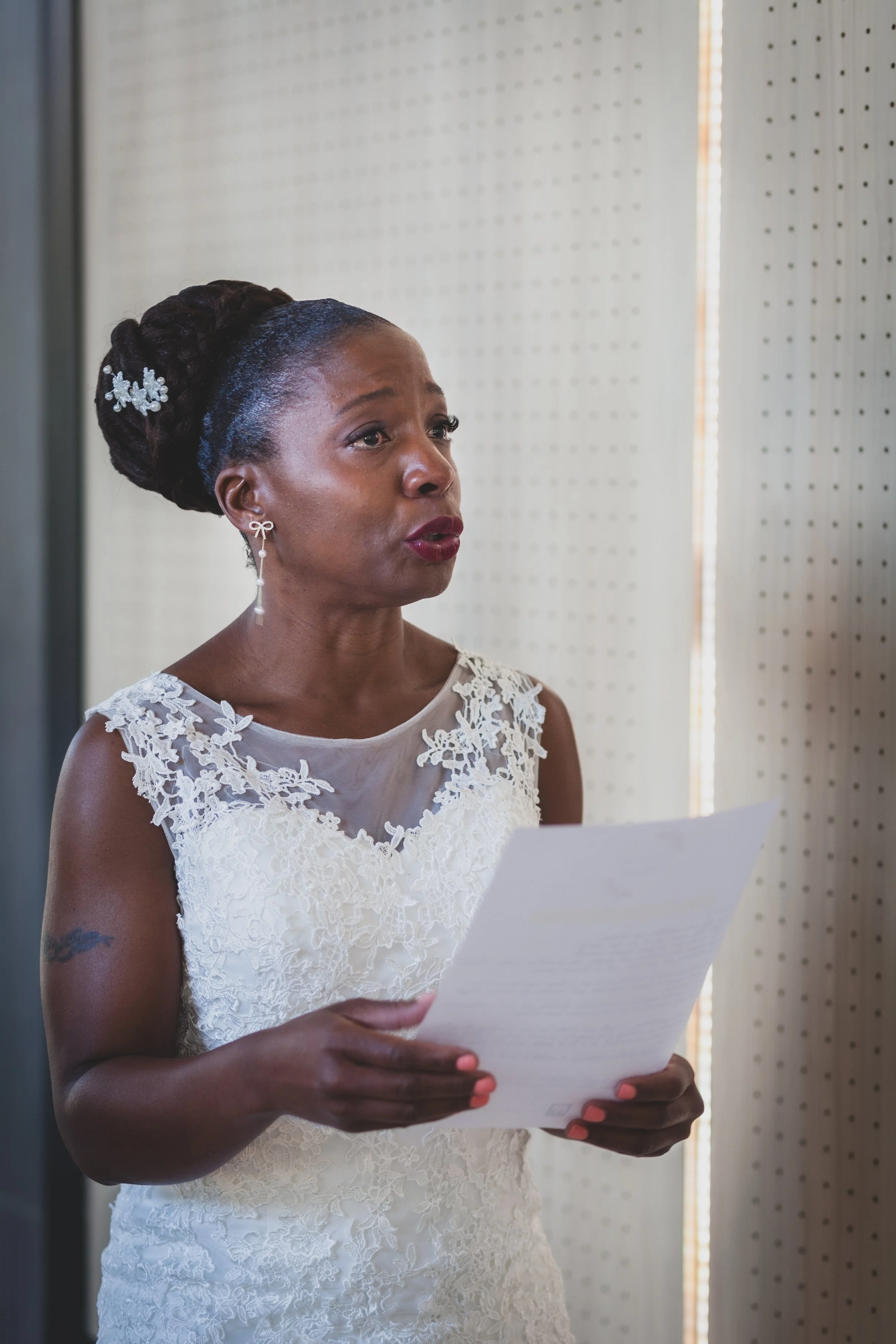 Une femme en robe de mariée lit un discours ou un texte, avec un fond clair et clair, cheveux tressés et accessoires élégants.