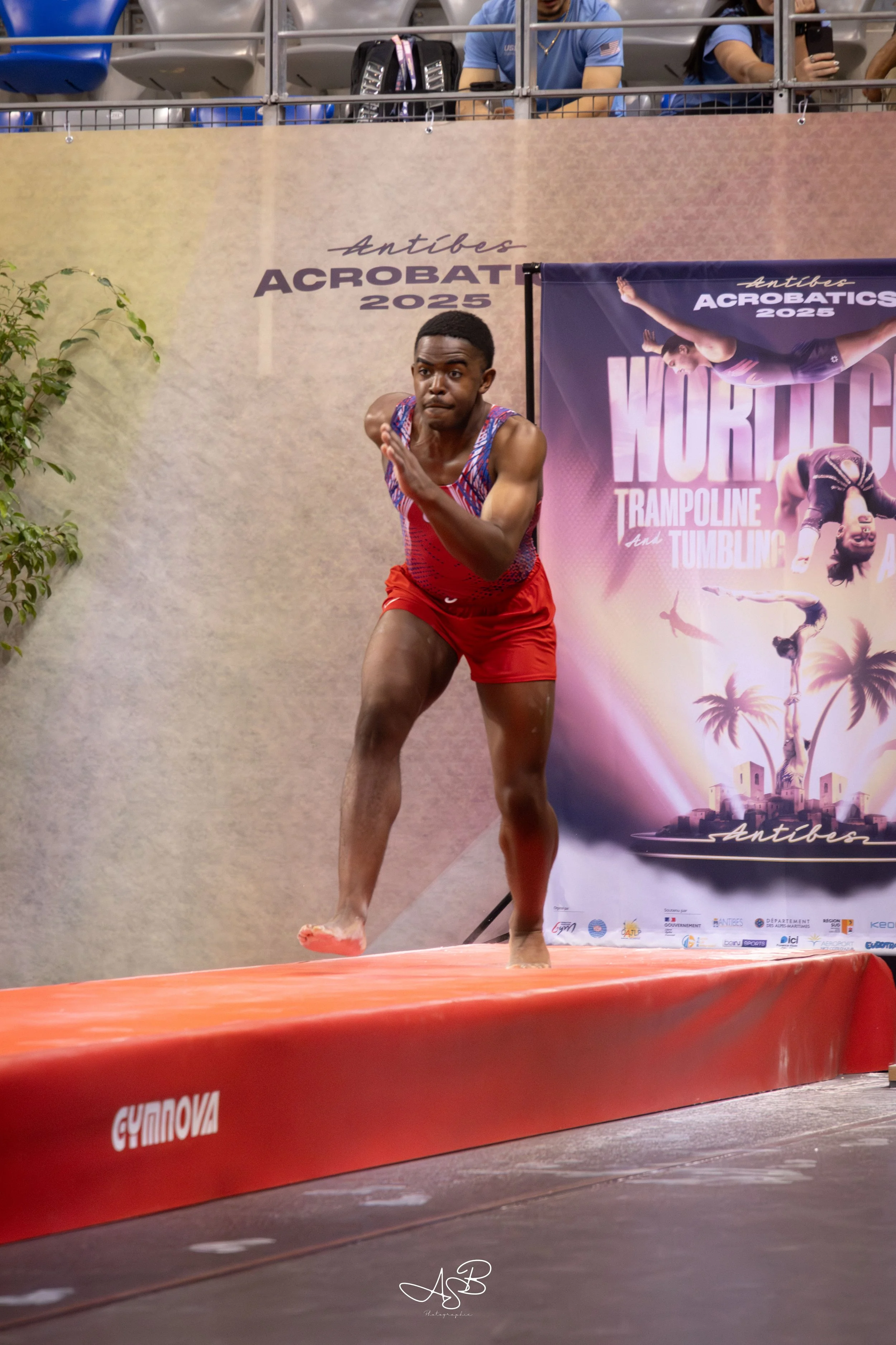 Un homme en tenue de gymnastique en mouvement lors d'une compétition d'acrogymnaste, sur une plateforme rouge, avec un panneau promotionnel en arrière-plan indiquant Antibes Acrobatic 2025 et le thème trapèze et tumbling.