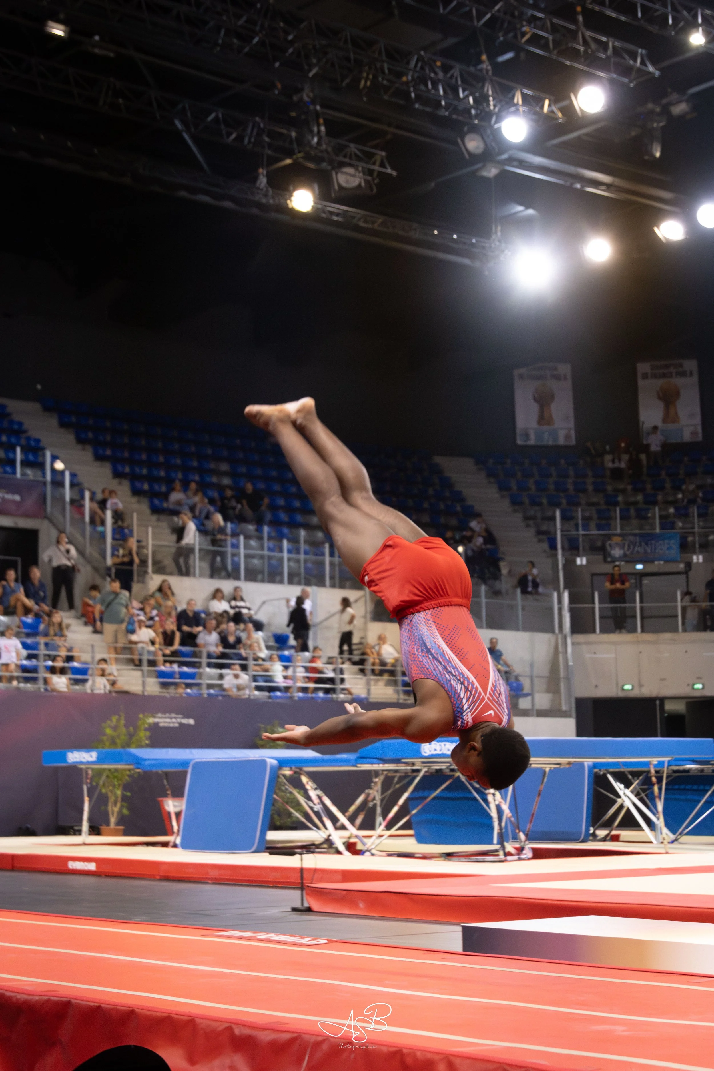 Une gymnaste effectuant un saut périlleux en gymnastique artistique lors d'une compétition, dans un gymnase avec des spectateurs en arrière-plan.