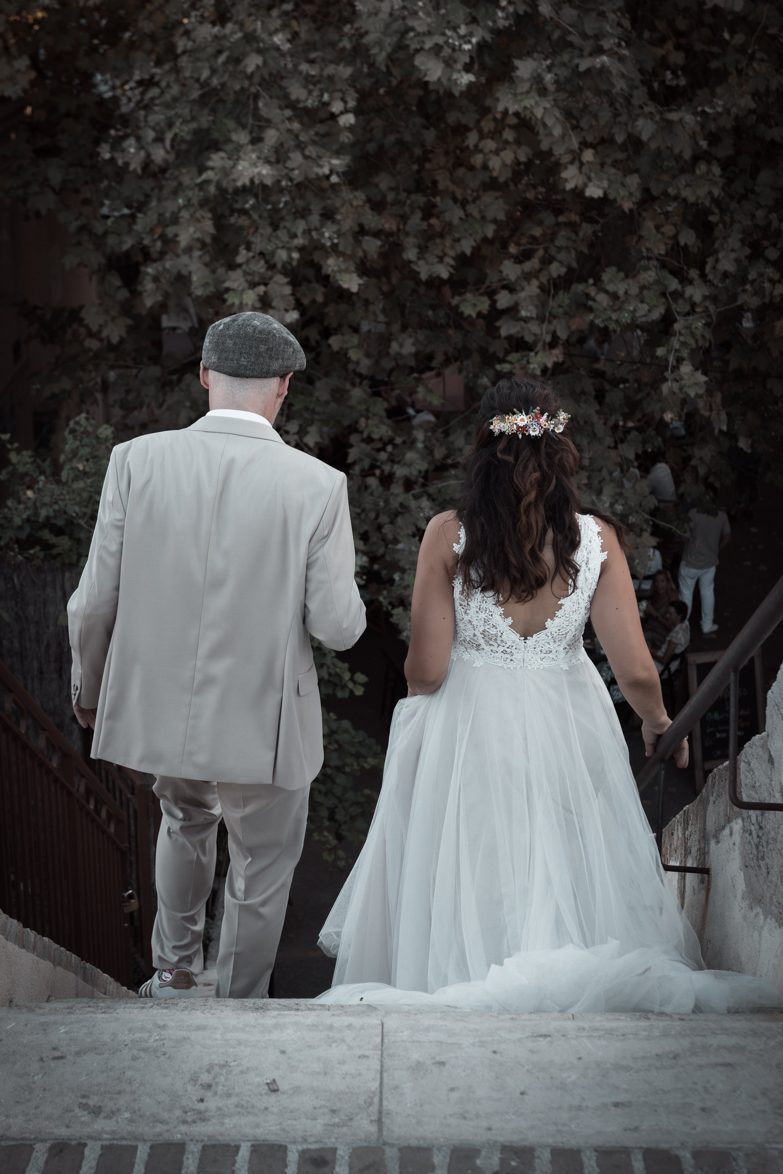 Un couple en tenue de mariage marche en haut d'un escalier en pierre, vus de dos, avec un arbre en arrière-plan.