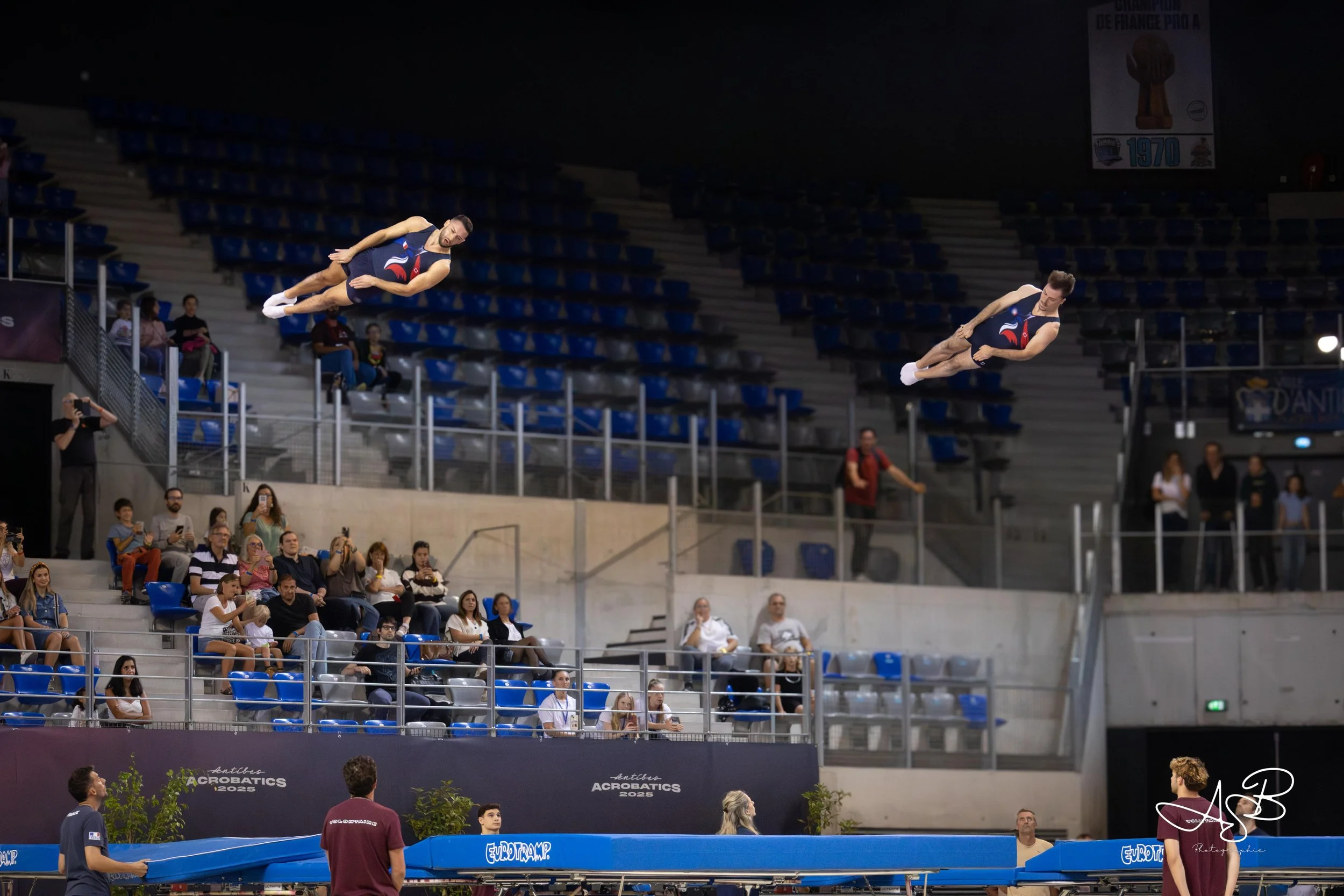Deux athlètes en train de faire de la gymnastique acrobatique en pleine volée lors d'une compétition, avec un public assis dans les gradins et quelques personnes prenants des photos.