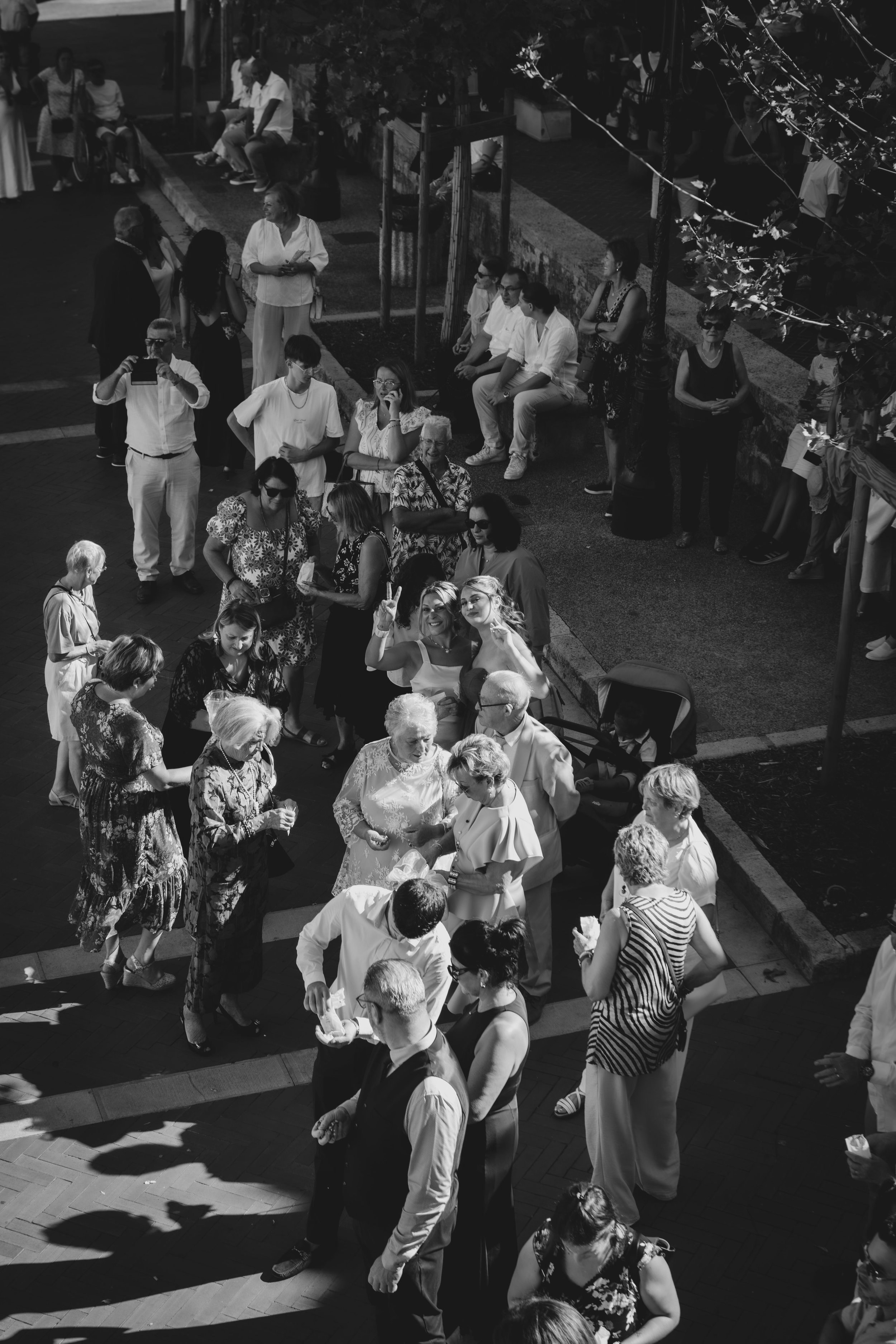 Groupe de personnes lors d'une réunion ou d'une fête en plein air, avec des personnes âgées, des jeunes, des adultes, certaines prennent des photos, d'autres discutent, certains sont assis et d'autres debout, sous un arbre avec des ombres et une ambi