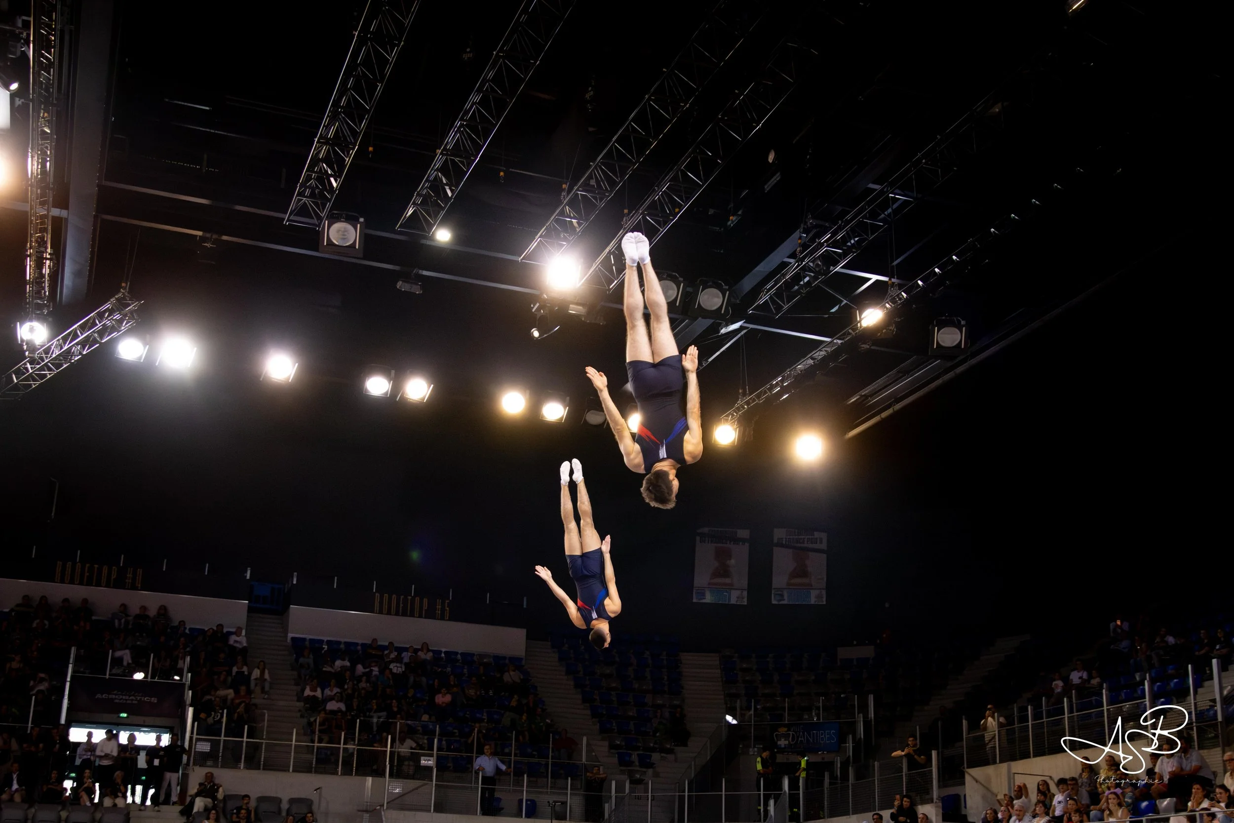 Deux gymnastes effectuant une acrobatie dans une salle de spectacle avec un public en arrière-plan, éclairée par de fortes lumières.