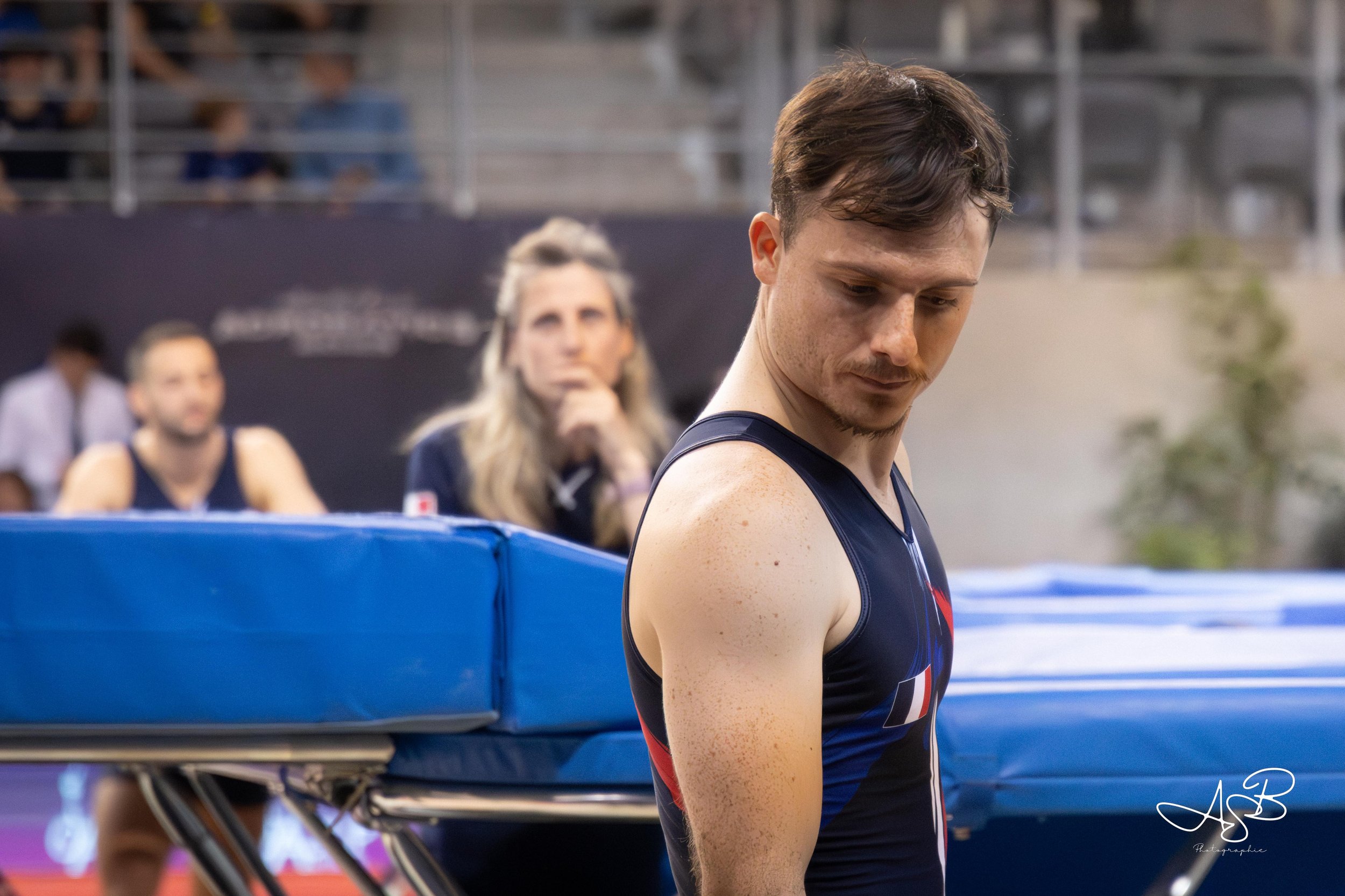 Un homme en tenue de sport regarde vers le sol dans un environnement d'athlétisme ou de saut en longueur, avec des spectateurs en arrière-plan.