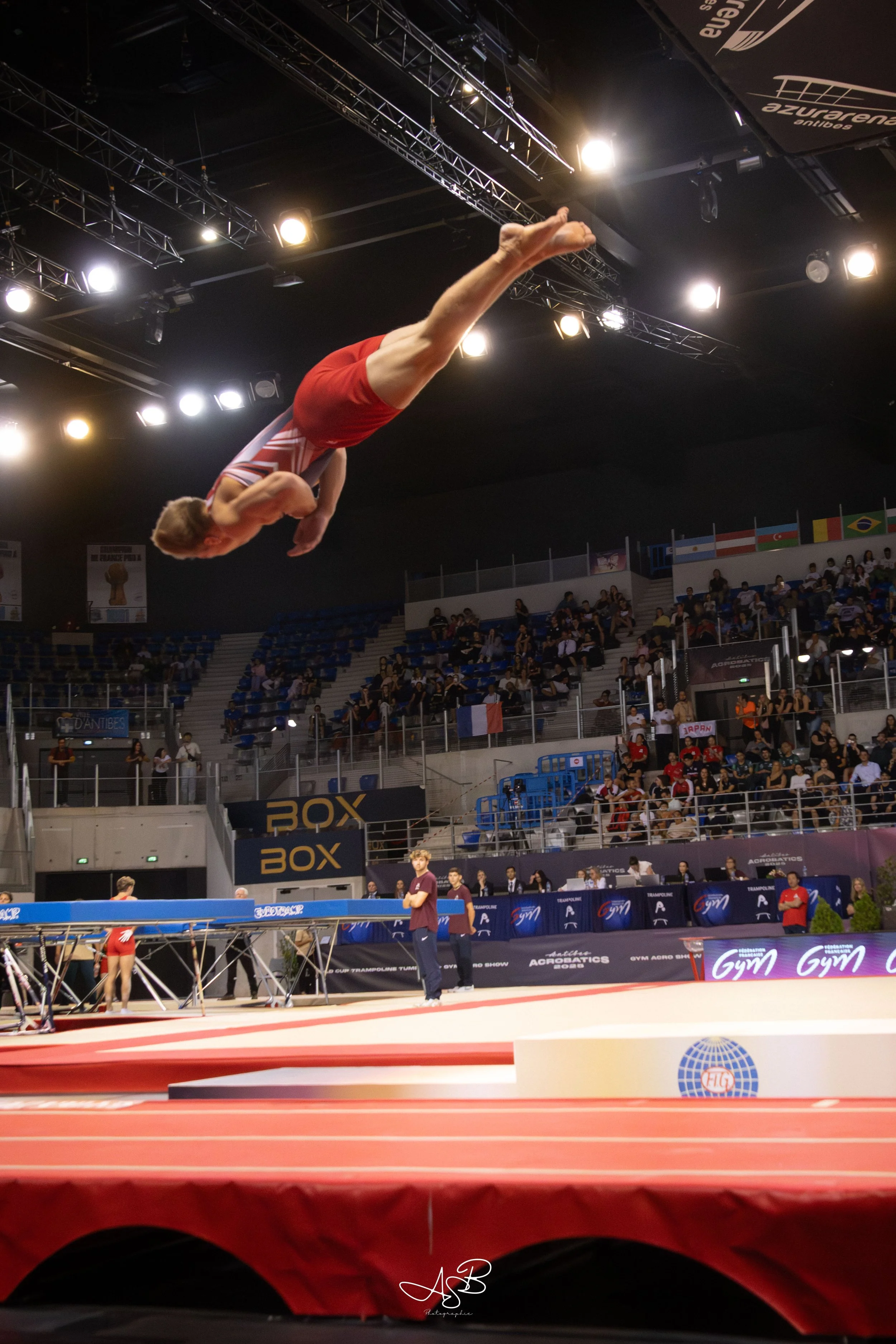 Un gymnaste en plein saut lors d'une compétition de gymnastique acrobatique, dans une arène avec des spectateurs et des drapeaux internationaux en arrière-plan.