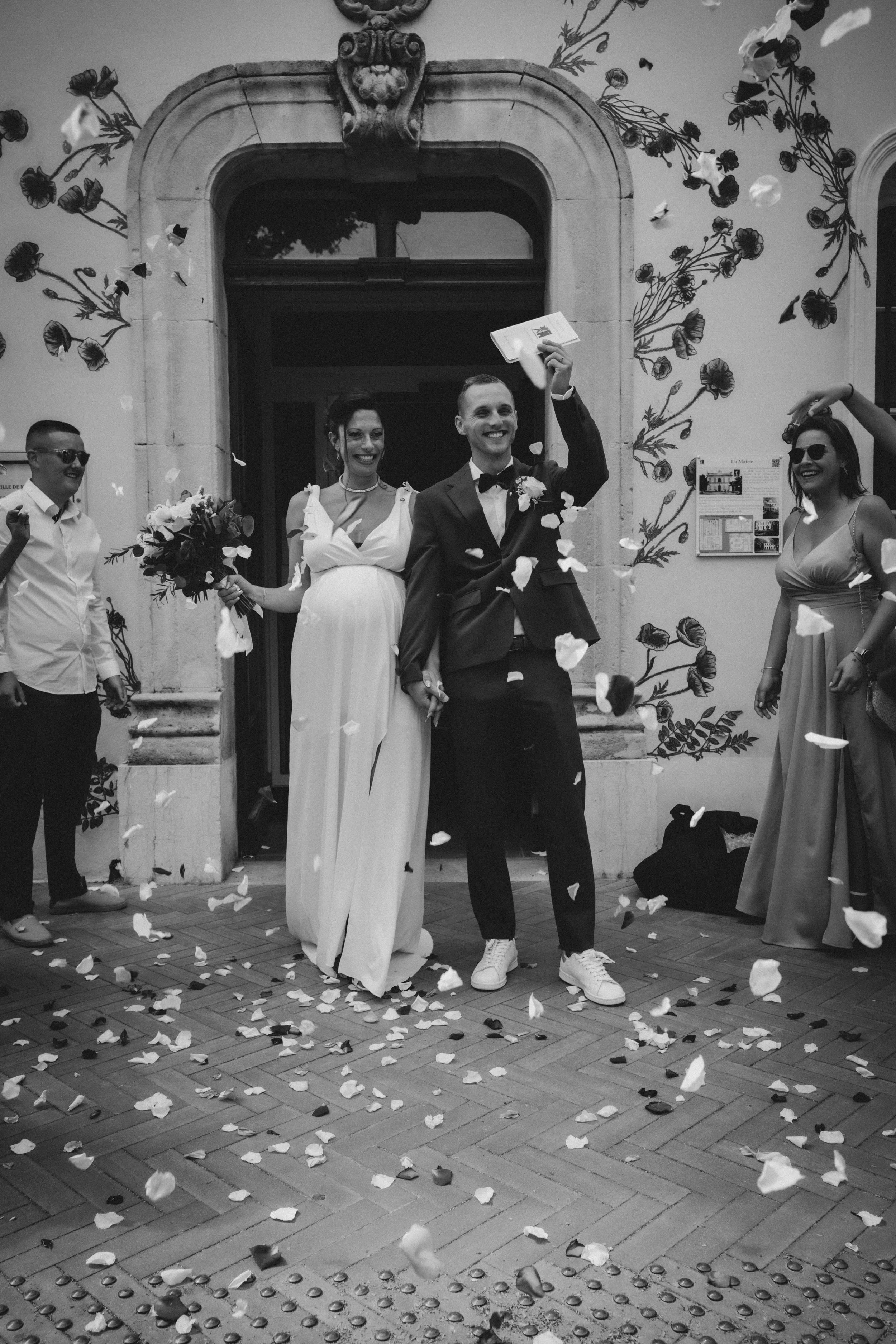 Un couple de mariés souriants sortant de l'église, entouré d'amis, avec des confettis, dans un style noir et blanc.