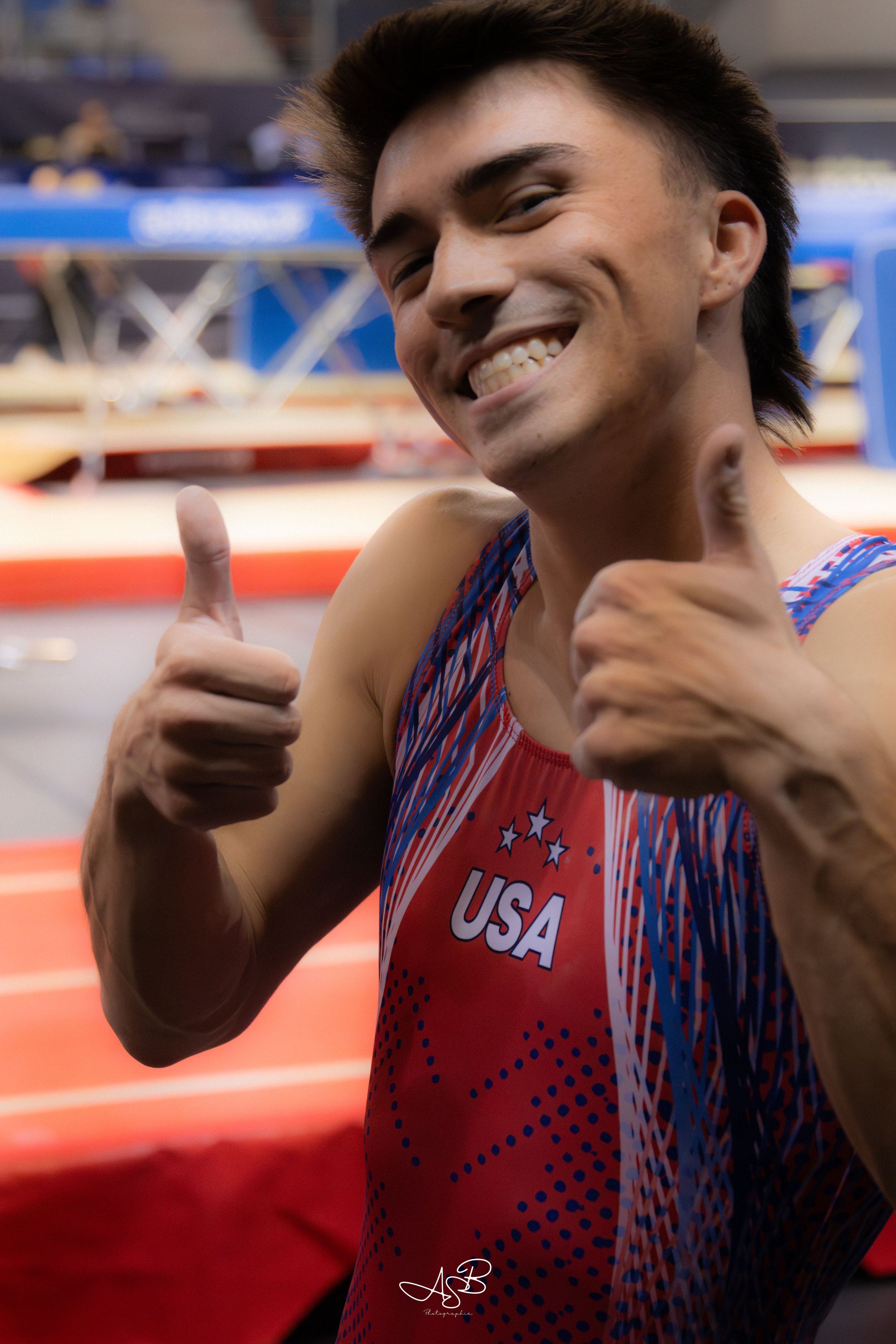 Un athlète américain souriant, portant un maillot rouge avec le mot USA, donnant un signe de pouce en l'air, dans un environnement d'athlétisme.