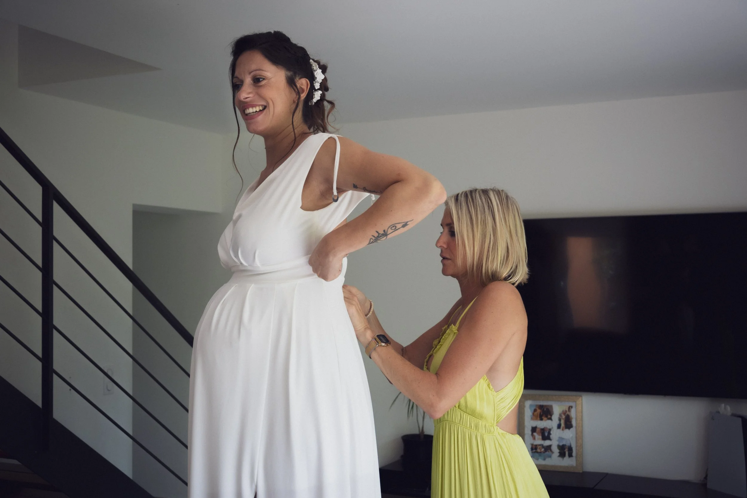 Deux femmes en train d'aider une femme à ajuster sa robe blanche de mariage à l'intérieur d'une maison.