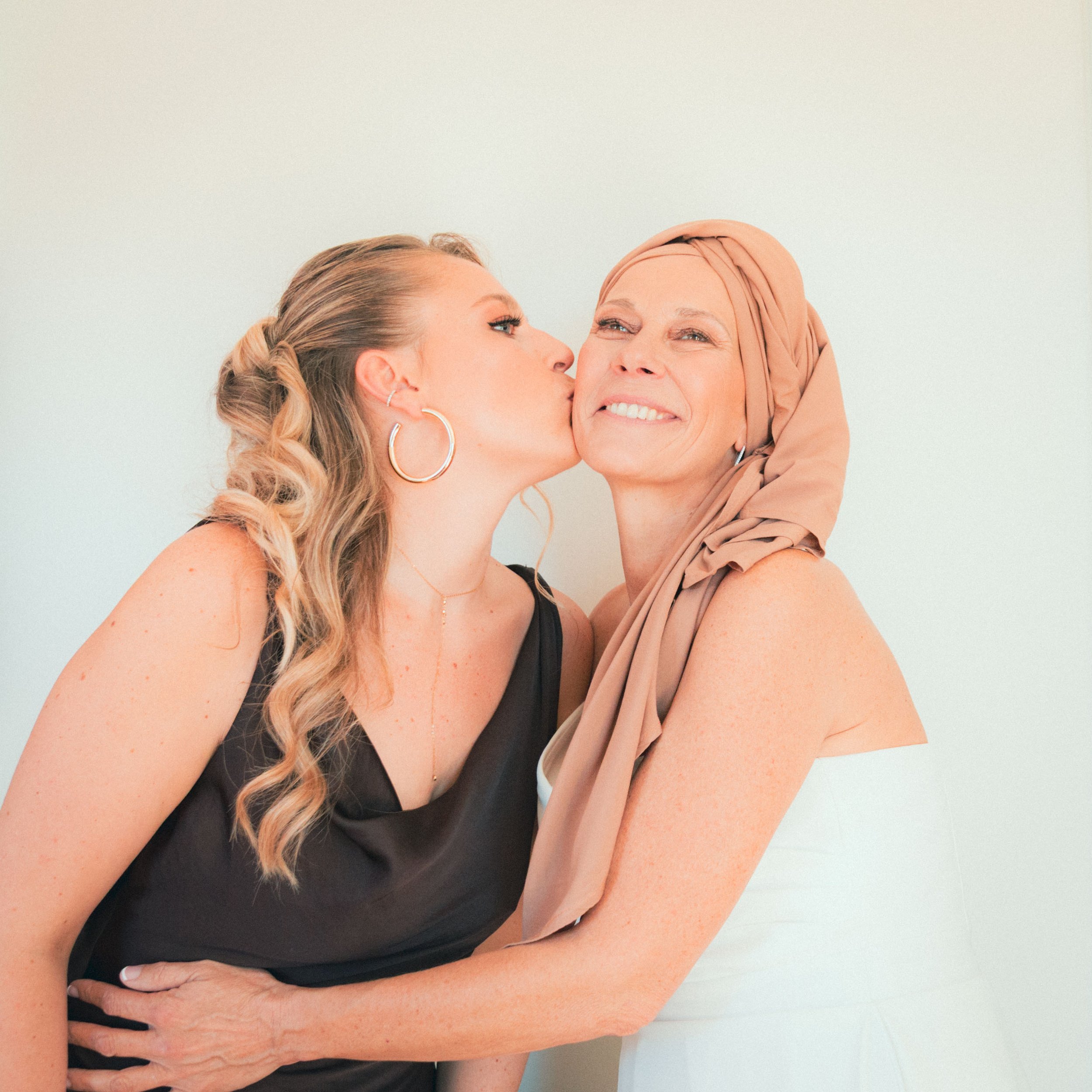 Deux femmes, une plus âgée avec un foulard beige, reçoit un baiser affectueux de la plus jeune, qui porte de grands anneaux d'oreilles et une robe noire, devant un mur blanc.