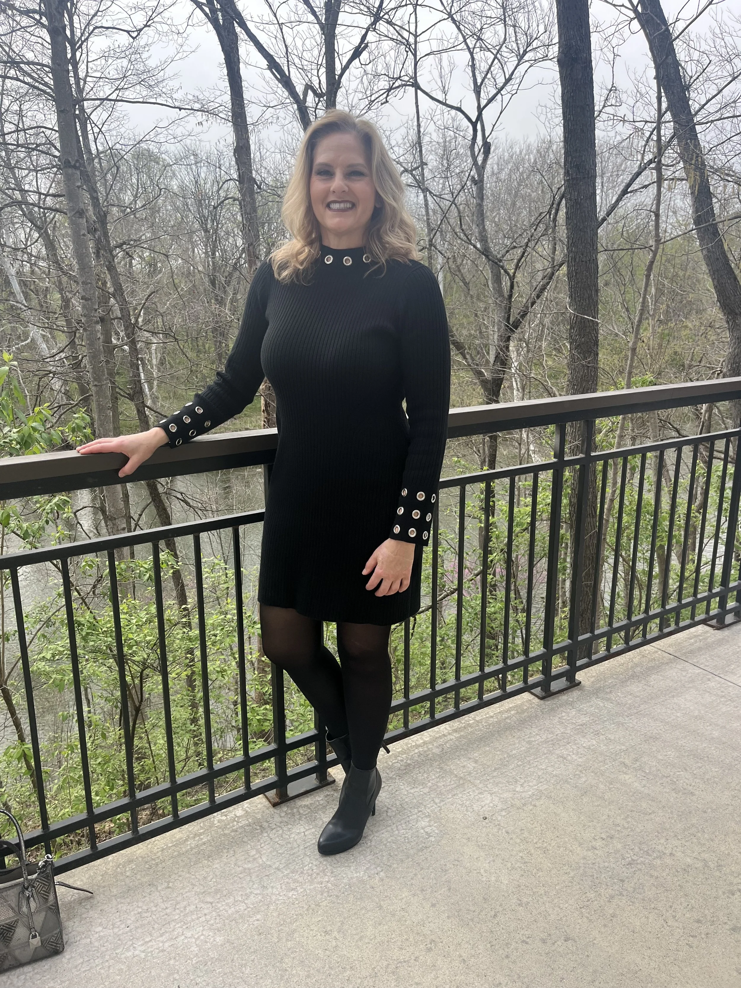 A woman with blonde hair in a black dress standing by a railing in front of woods and a creek
