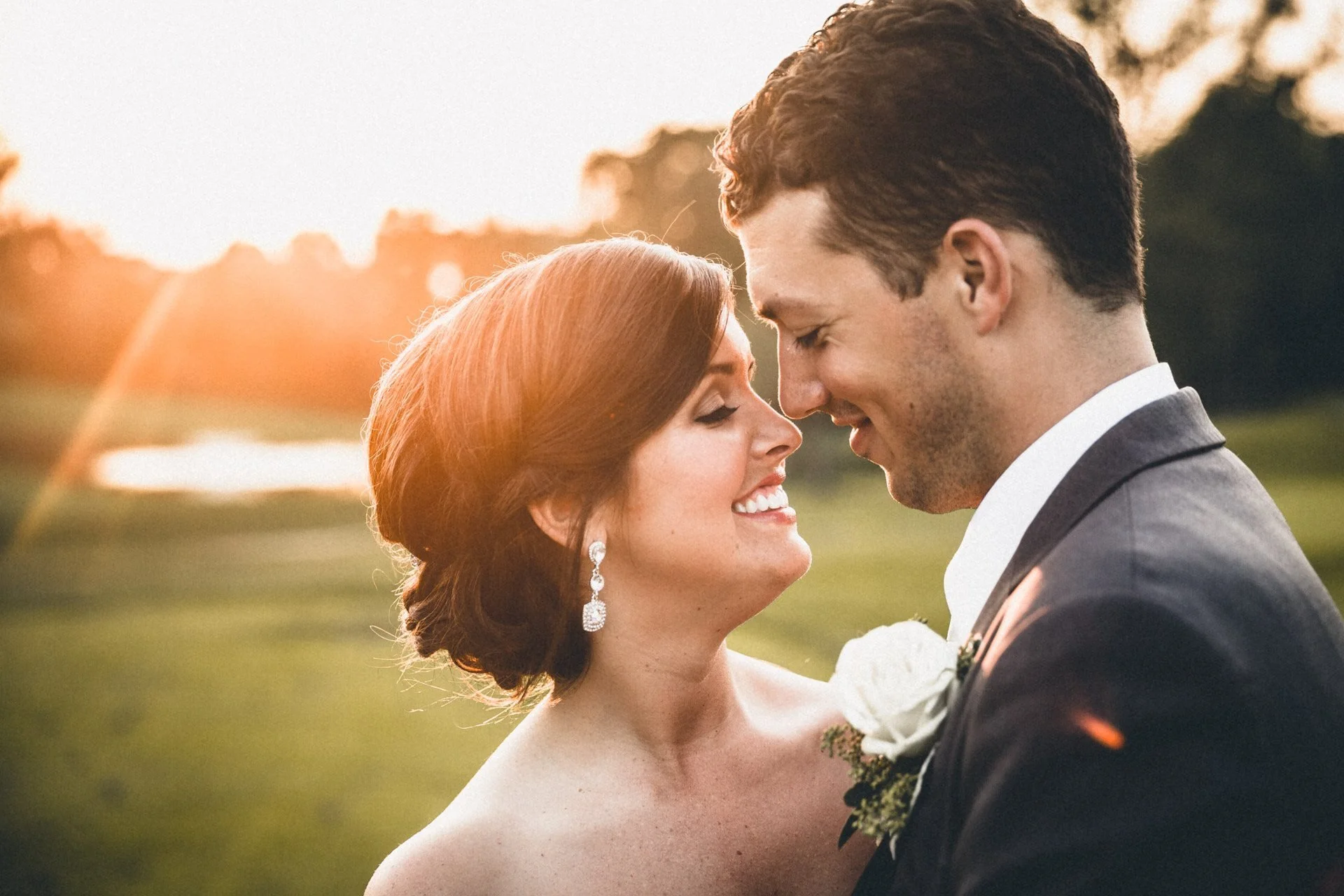 A smiling bride and groom with foreheads touching outdoors during sunset.