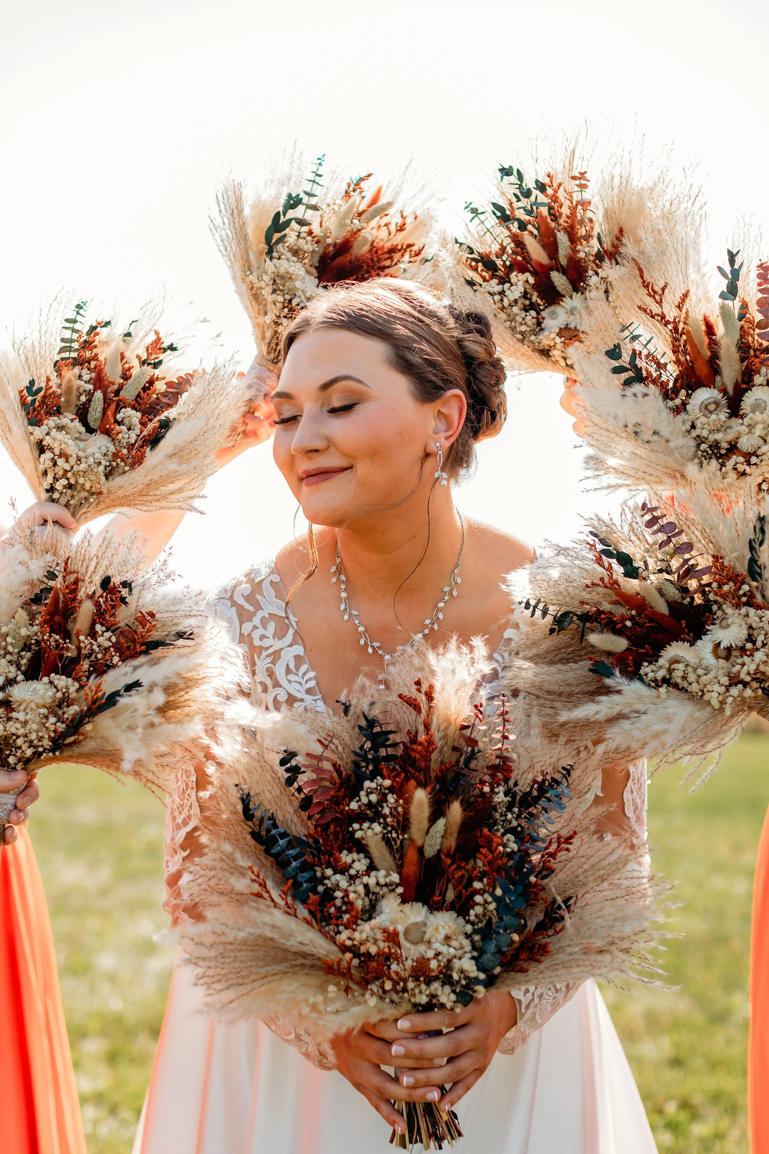 Bridal Makeup Look by Colour By Courtney Smiling woman in a wedding dress, holding a large floral bouquet surrounded by matching floral arrangements, outdoors on a sunny day.