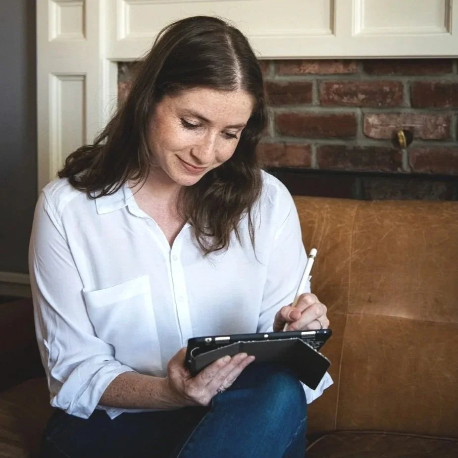 A woman with brown hair, wearing a white shirt, sitting on a brown leather couch, writing in a notebook with a pen.