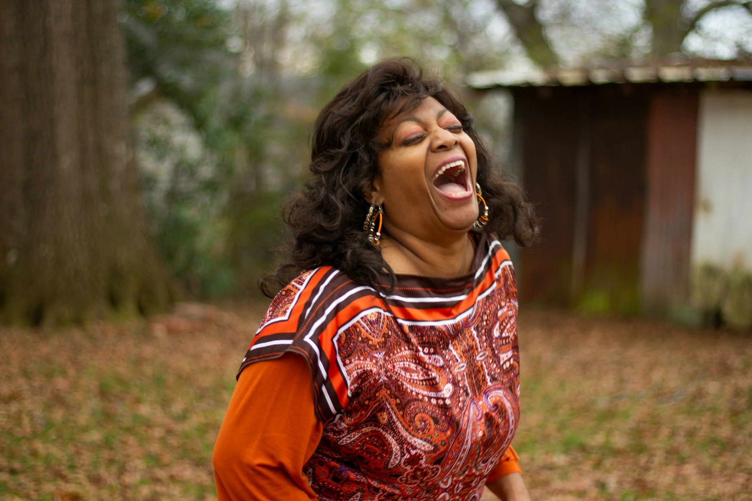 A woman with curly dark hair laughing outdoors, wearing a colorful patterned top and orange sleeves, in a park with trees and a wooden shed in the background.